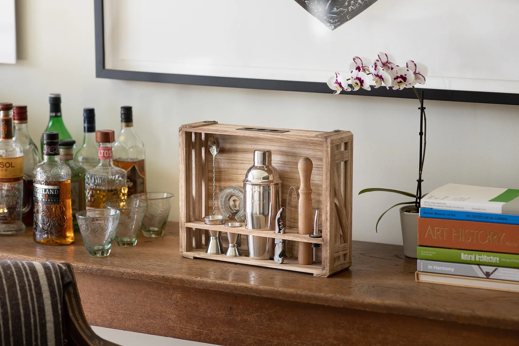 A wooden bar cart holding cocktail tools, and barware with liquor bottles and glasses on a wooden table, with books and a potted orchid plant in the background.