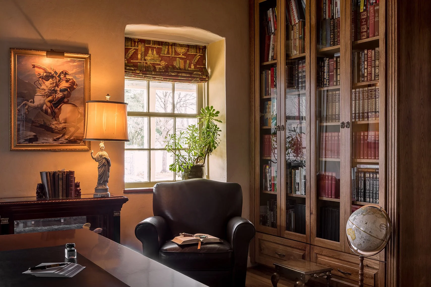 A cozy study room with a window, a bookshelf filled with books, a leather armchair, a globe, and a painting of a man on a horse on the wall.