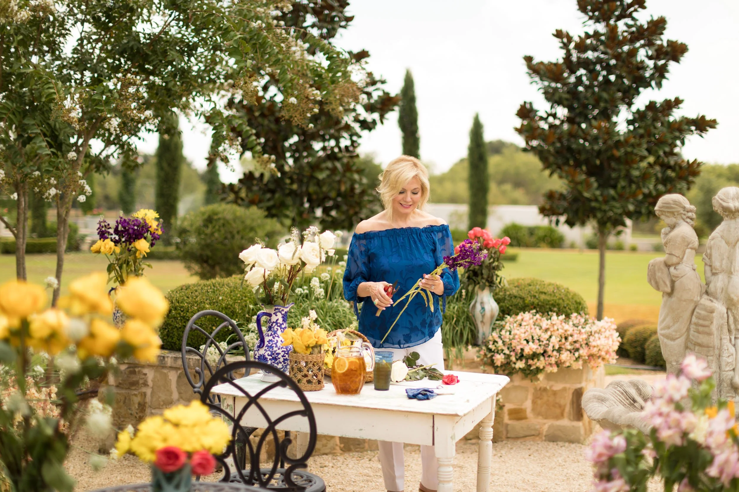 A woman with blonde hair in a blue off-shoulder dress arranging flowers at a table outdoors. The table has various vases with colorful flowers, and there are sculptures and garden trees in the background.