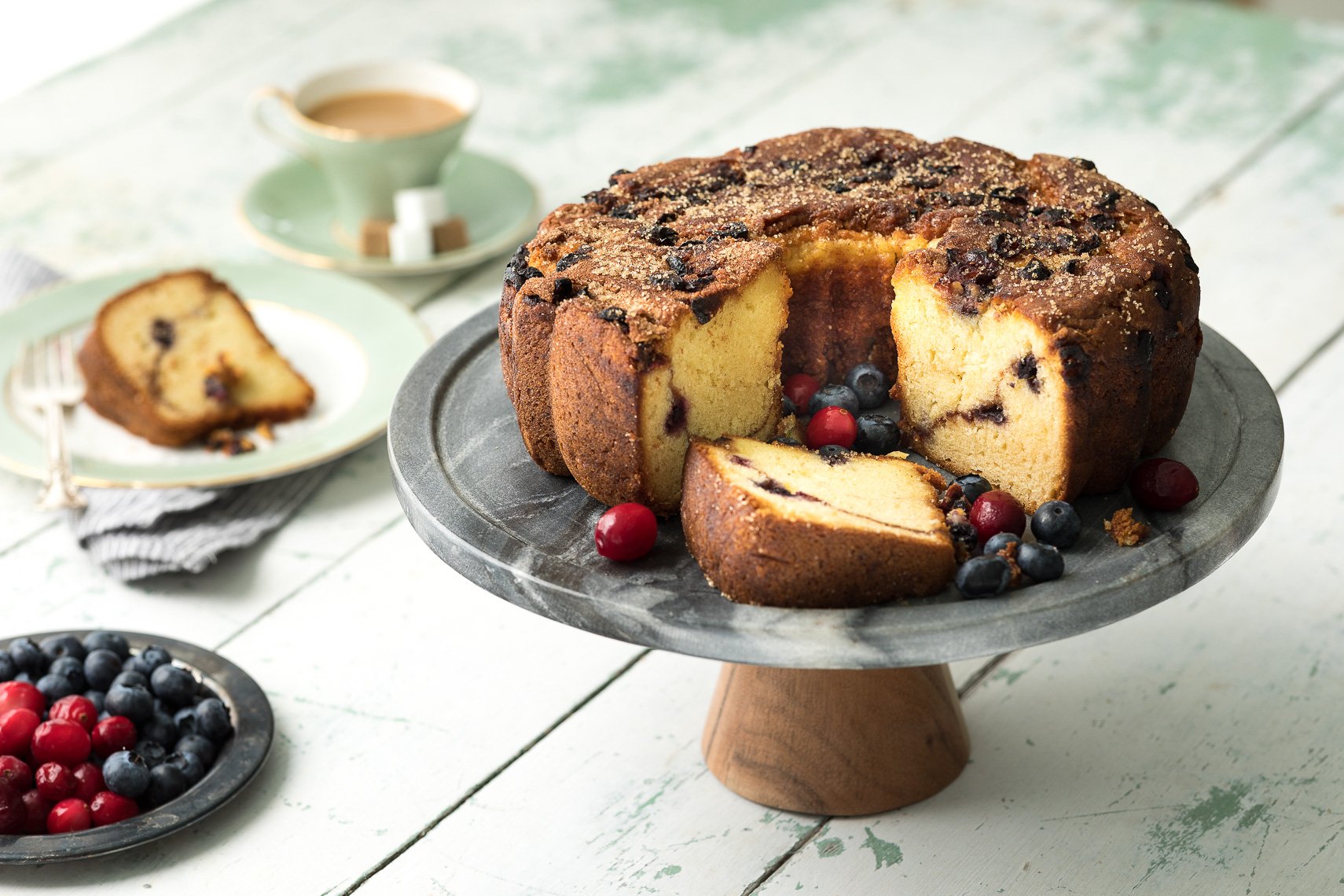 Bundt cake with blueberries and cranberries, partially sliced on a cake stand, with a cup of coffee and a plate of cake slices in the background, on a rustic white wooden table.