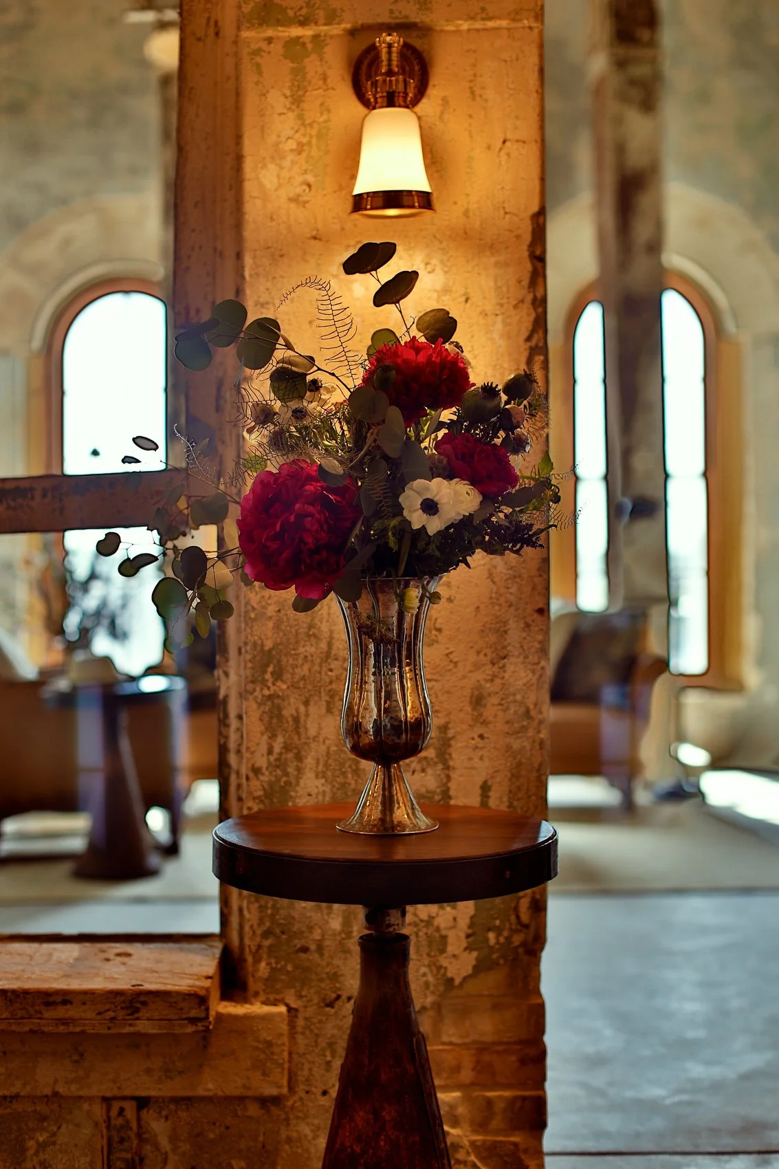 A glass vase with a bouquet of red, white, and purple flowers, along with green foliage, on a round wooden table in a warmly lit interior with rustic walls and arched windows.