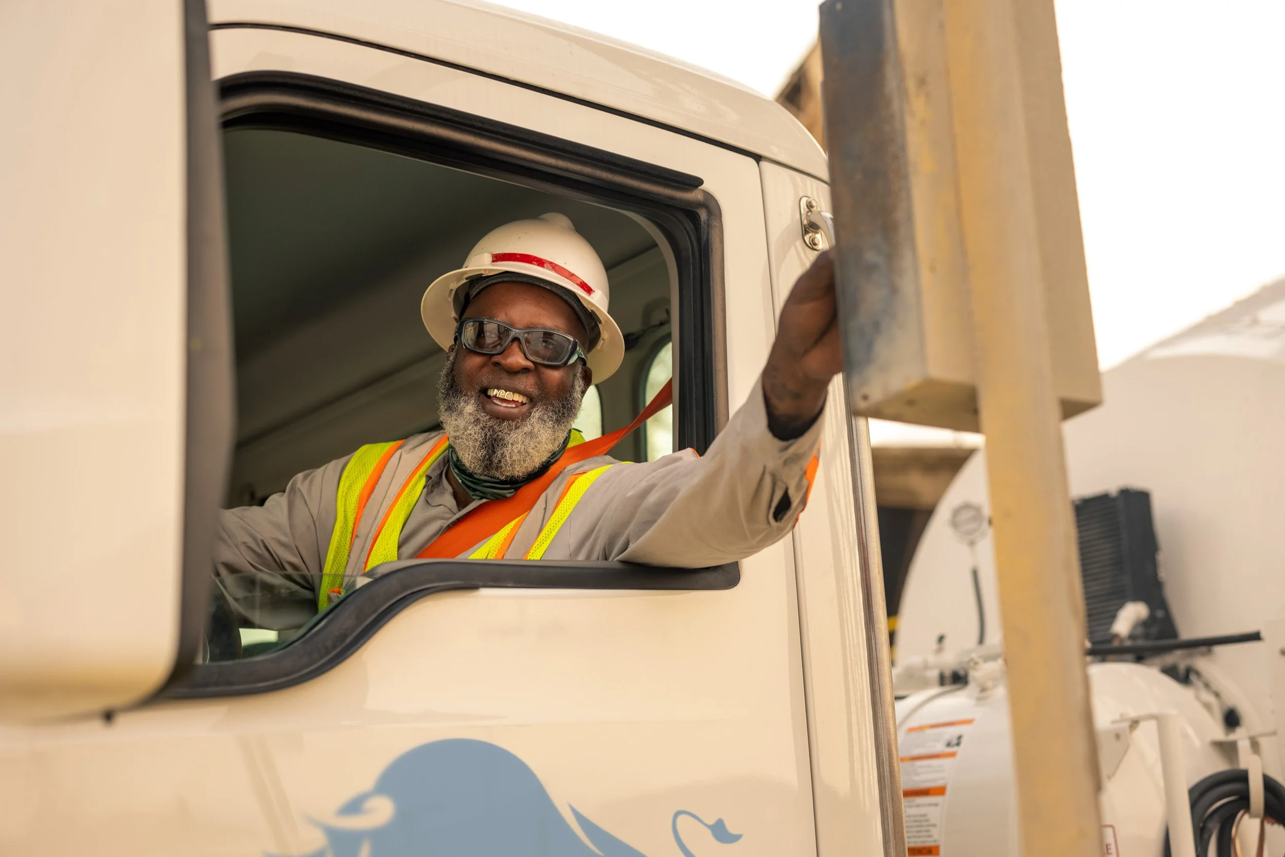 A smiling man with a beard, wearing sunglasses, a white hard hat with a red stripe, and safety vest, is leaning out of a truck window and waving.