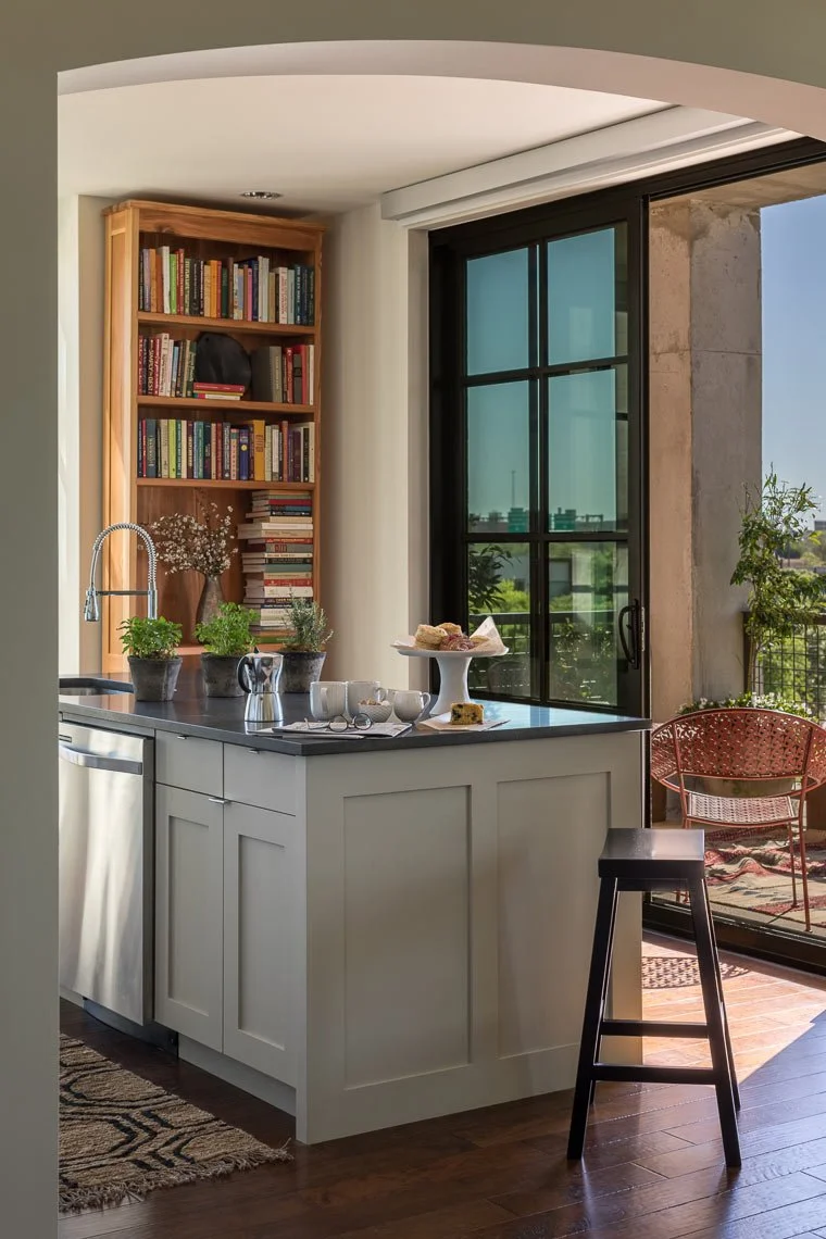 Kitchen with white cabinets, a black countertop, potted plants, a wet bar with a teapot and cups, a stack of books, and a sliding glass door leading to a balcony with outdoor furniture.