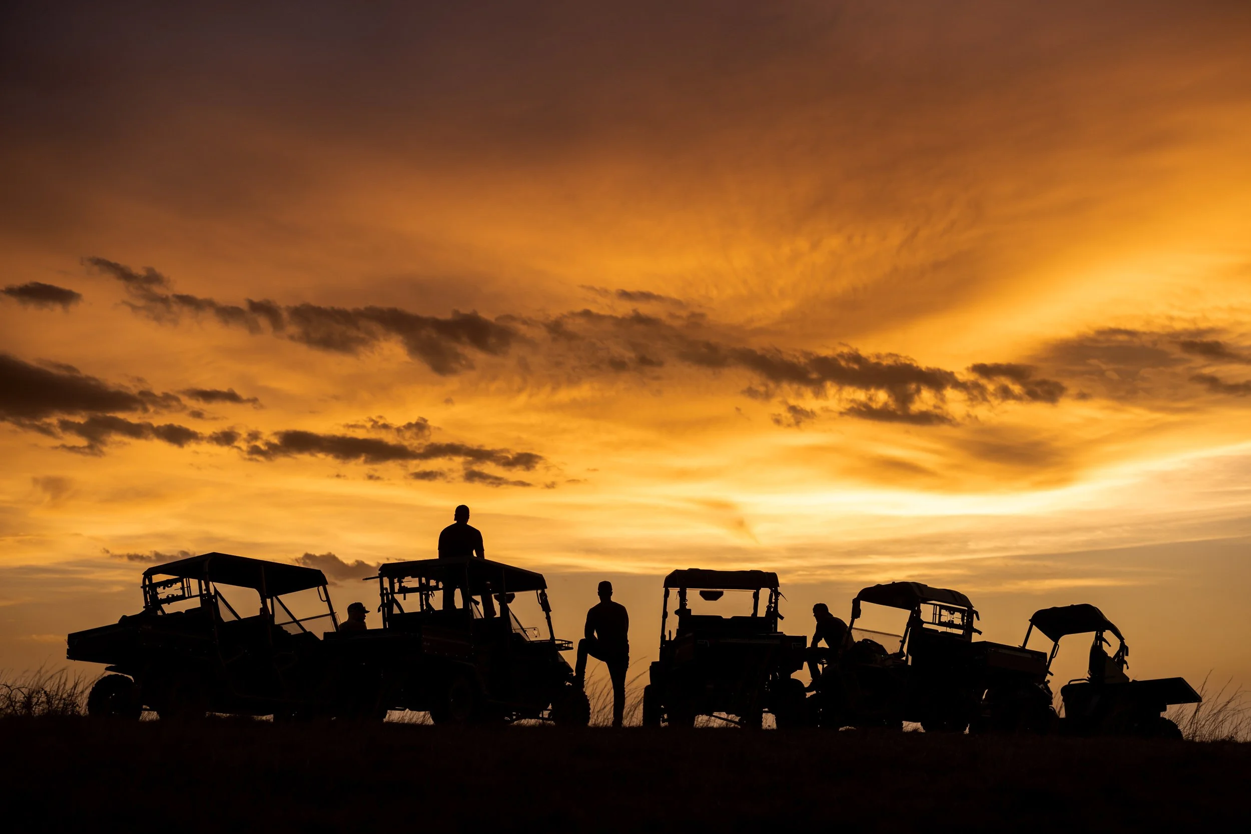 Silhouettes of people near boats during a vibrant orange sunset with a dramatic sky and scattered clouds.