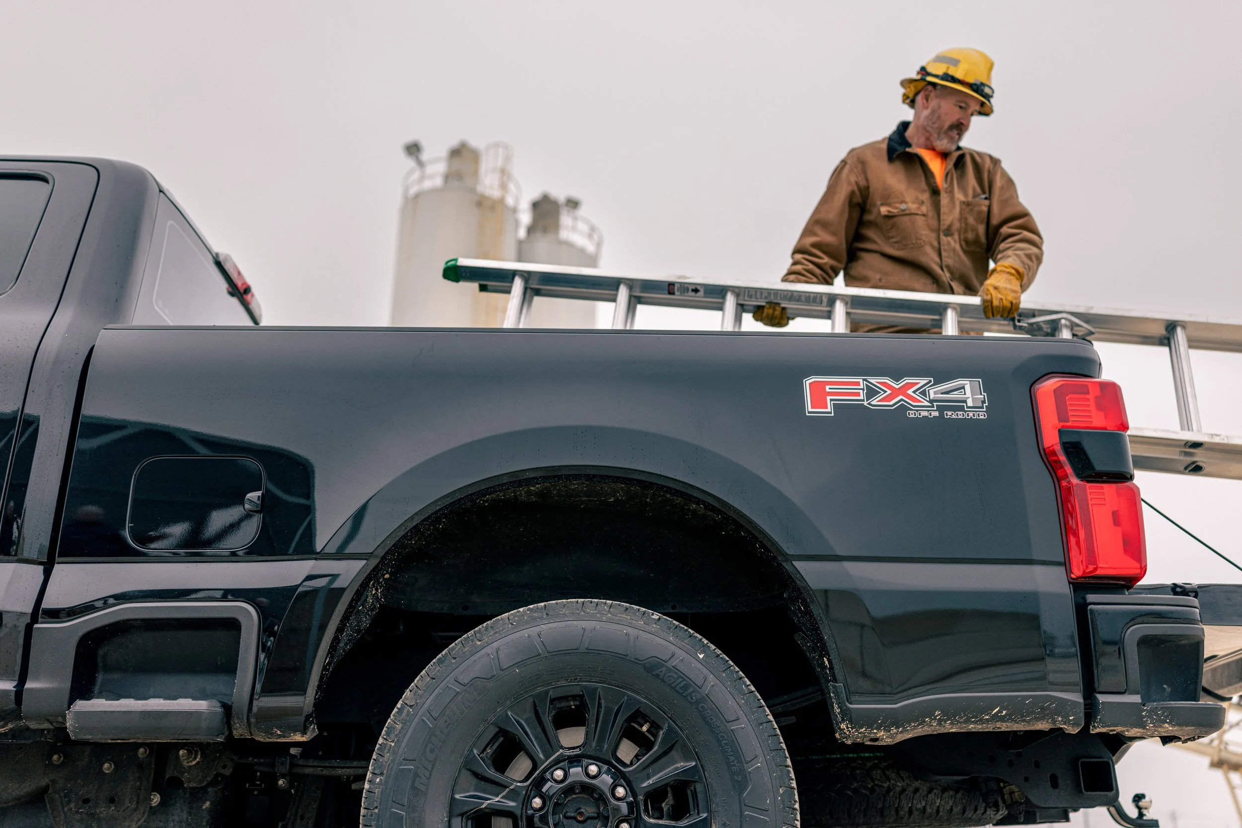 A man wearing a yellow safety helmet, brown work jacket, and yellow gloves standing on the back of a black pickup truck with a ladder, against a cloudy sky and industrial storage tanks in the background.