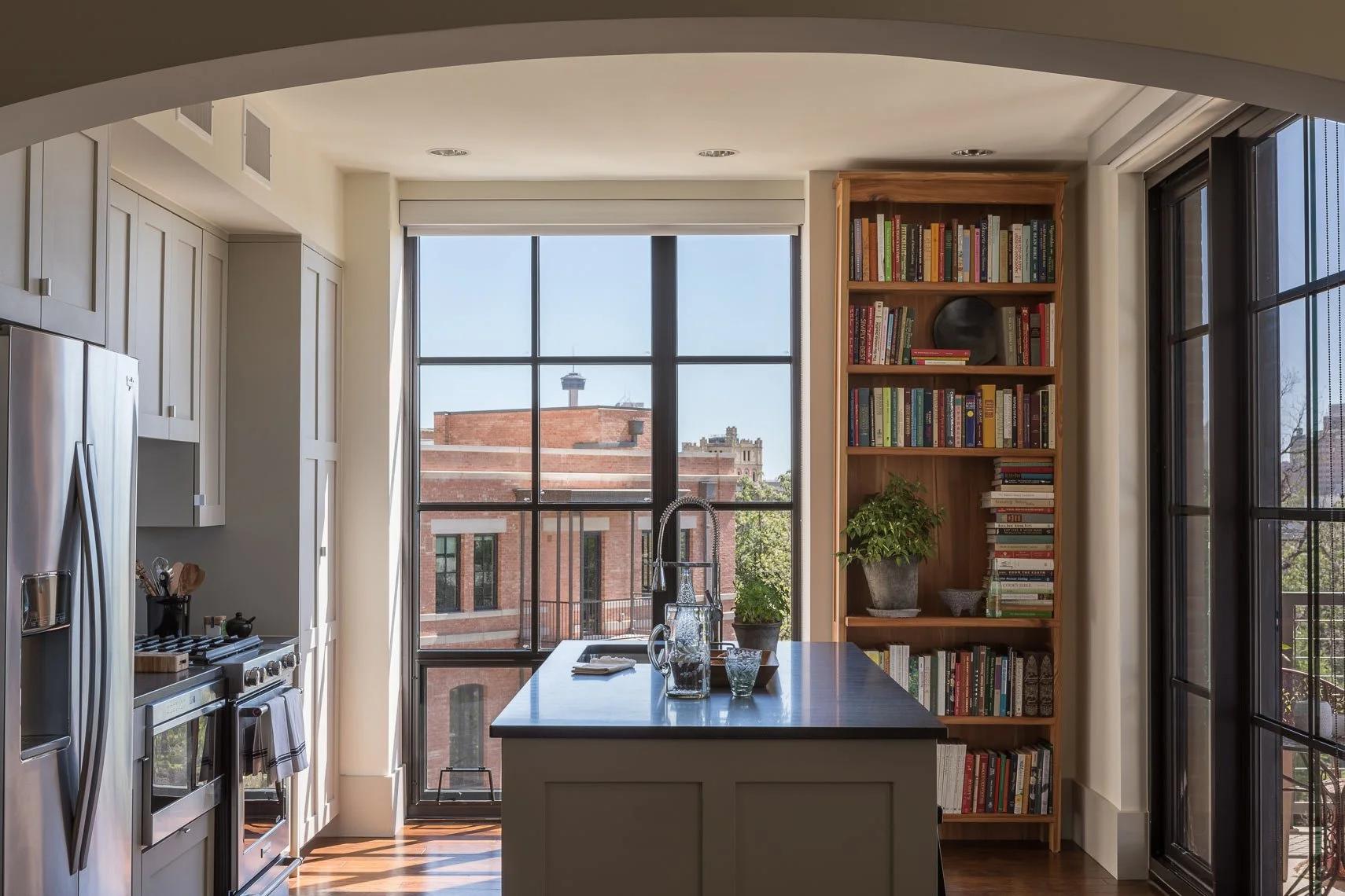 Modern kitchen with large windows, stainless steel refrigerator, oven, kitchen island with glassware and a sink, shelves with books, and potted plants with city buildings outside.