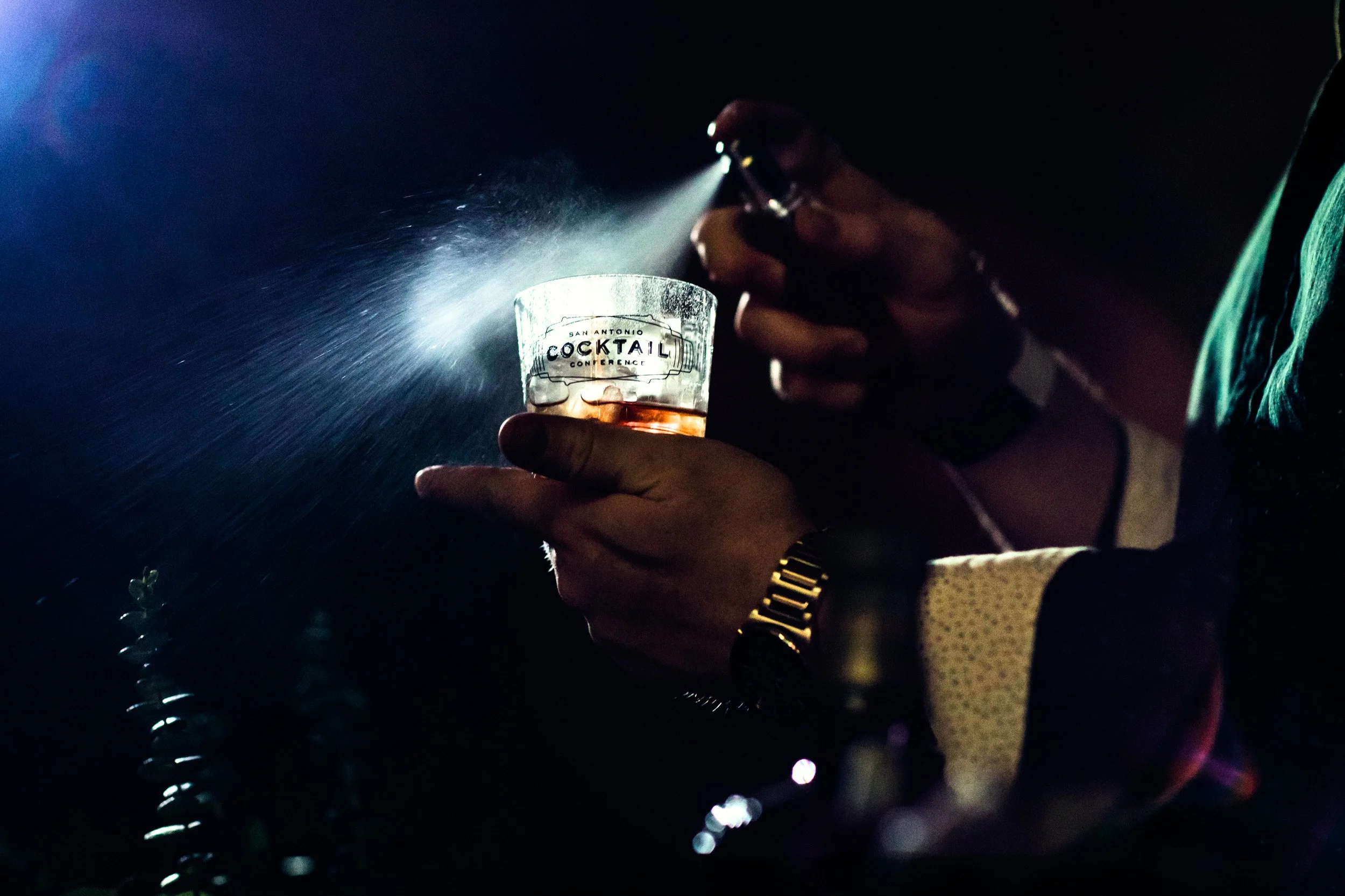 A person holding a glass with the words 'San Antonio Cocktail Conference' written on it, while using a small flashlight to illuminate the glass in a dark setting.