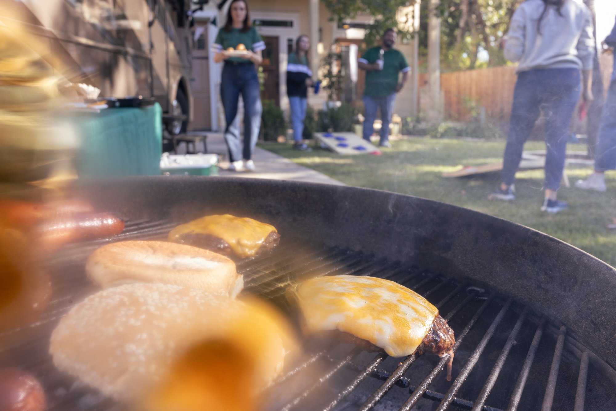 Close-up of cheeseburgers cooking on a grill, with a backyard gathering in the background. People are standing and socializing near a house, some holding plates of food.