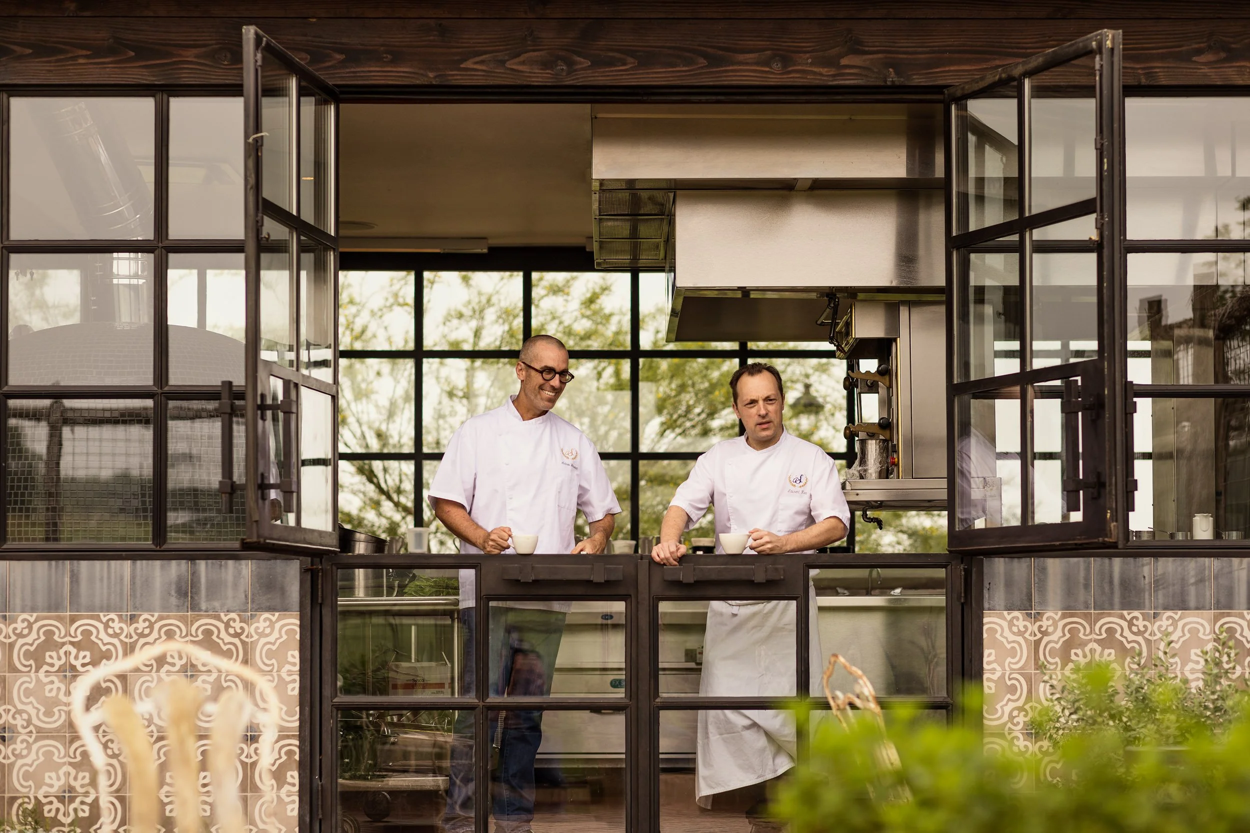 Two male chefs in white uniforms and glasses stand inside a modern kitchen with large windows, holding bowls, and smiling as they prepare food.