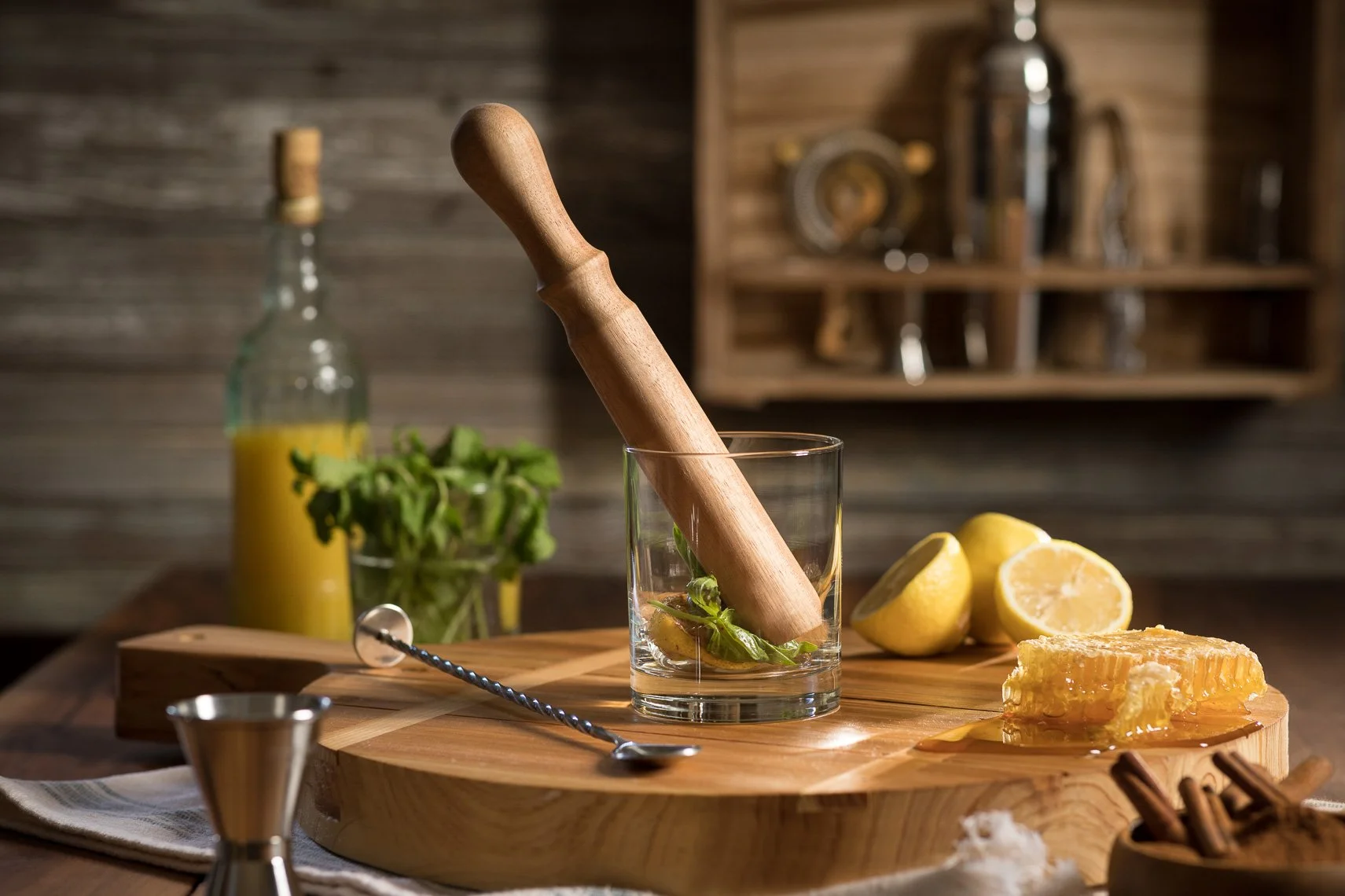 A wooden mortar and pestle in a glass, filled with lemon slices and fresh mint. There are lemons cut in half, honeycomb, a small honey jar, a bottle of yellow liquid, and a bowl of cinnamon sticks on a wooden cutting board. The background features ru