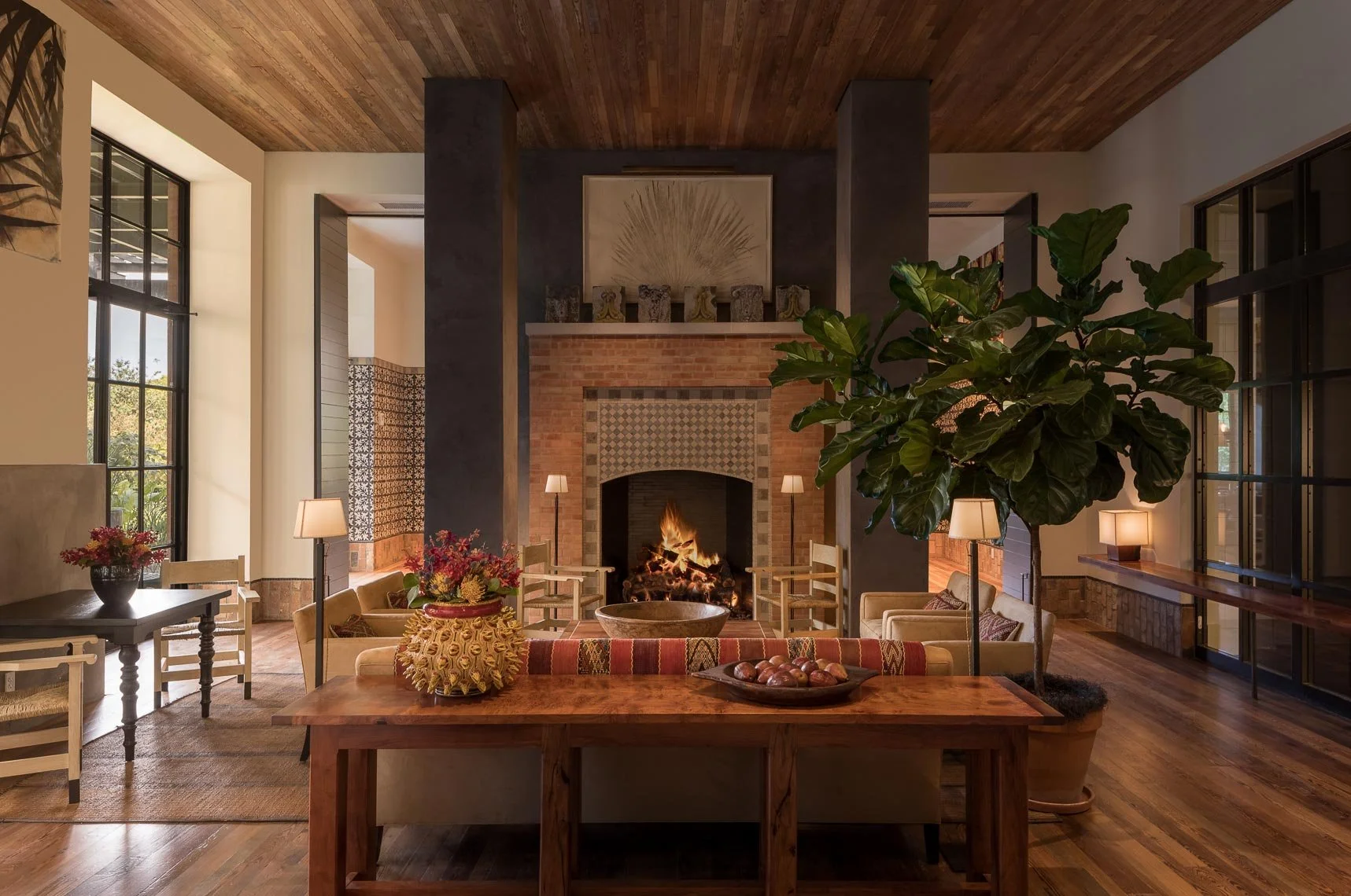 Living room with a brick fireplace, surrounded by sofas, chairs, and tables, a large indoor plant, and several lamps, with natural light coming through large windows.