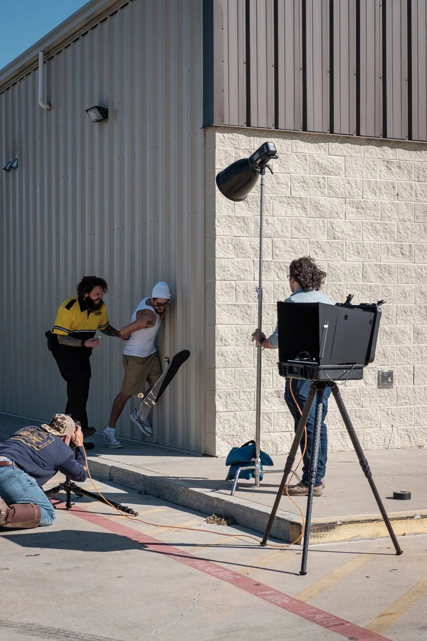 Photographers film a skateboarder pushing against a wall outside a building, with studio lights set up nearby.