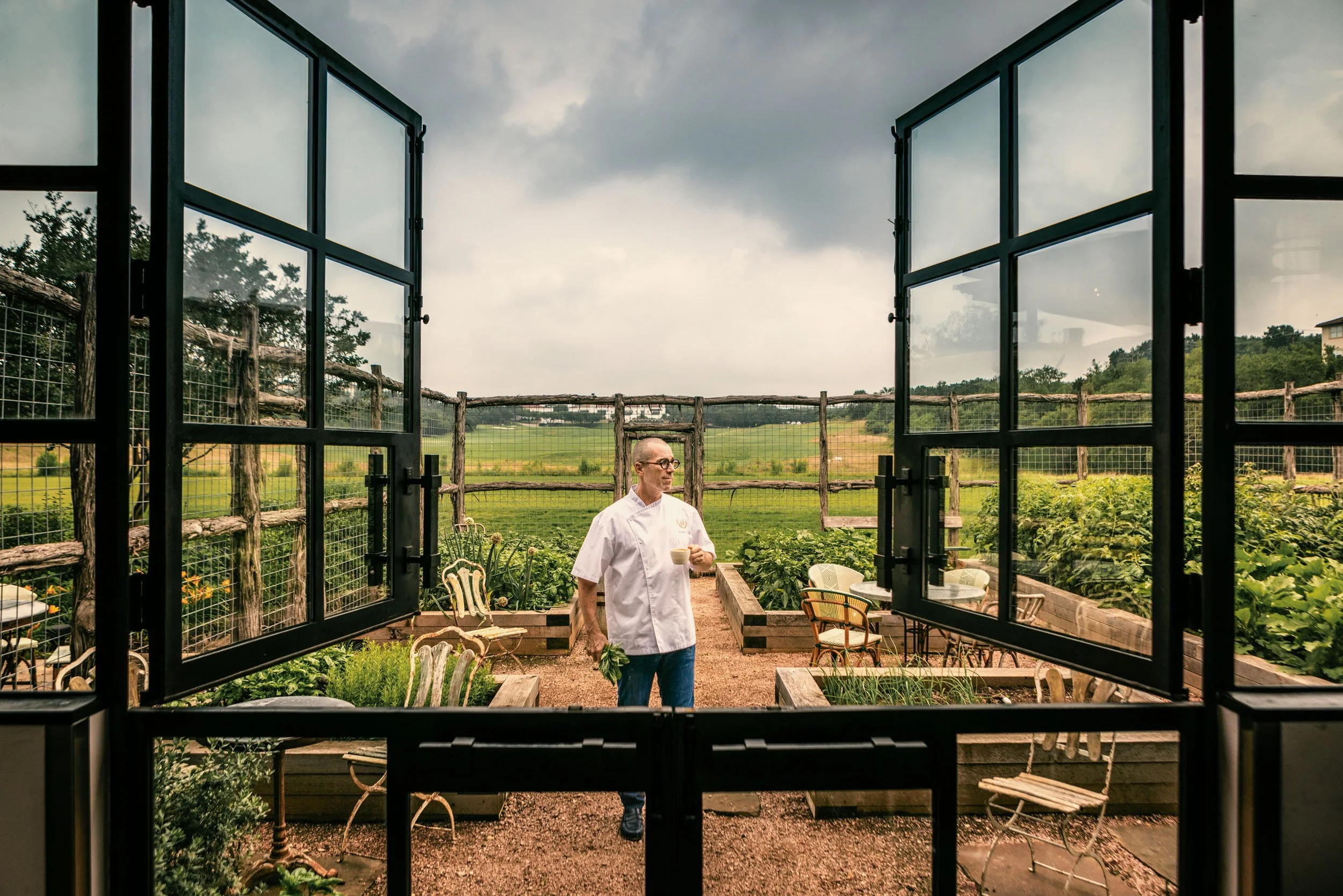 A man in a white chef's coat holding a cup in a garden cafe, viewed from an open window with a rustic outdoor landscape featuring a cloudy sky, wooden fencing, and green fields.