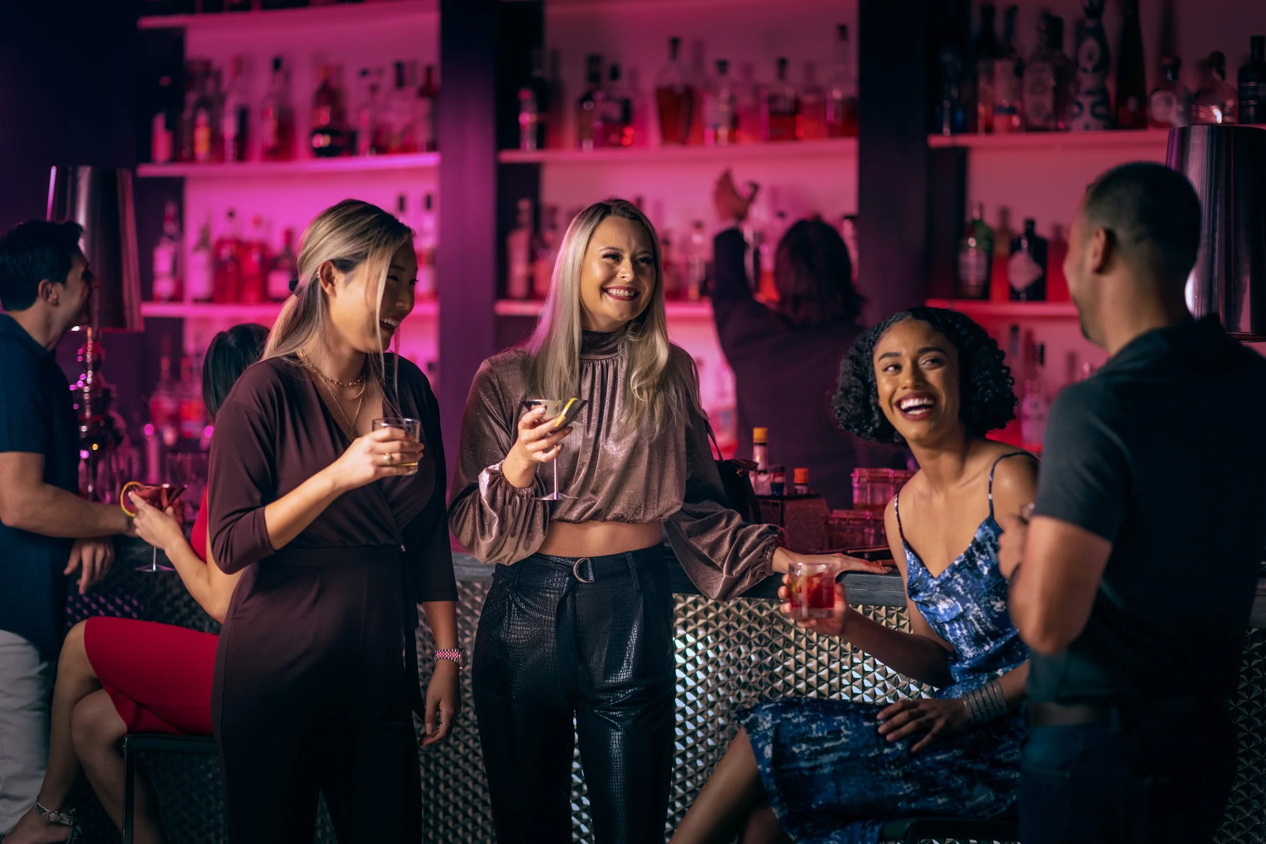 A group of friends at a bar, two women smiling and talking, holding drinks. The bar has pink and purple lighting, with bottles on shelves in the background.