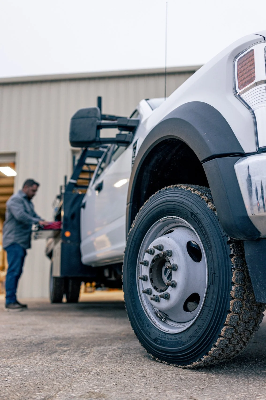 A man working on a white pickup truck inside a garage or workshop. The truck's front tire is close to the camera, with visible dirt and tire treads. The man appears to be checking or working on the vehicle near a tool cart.