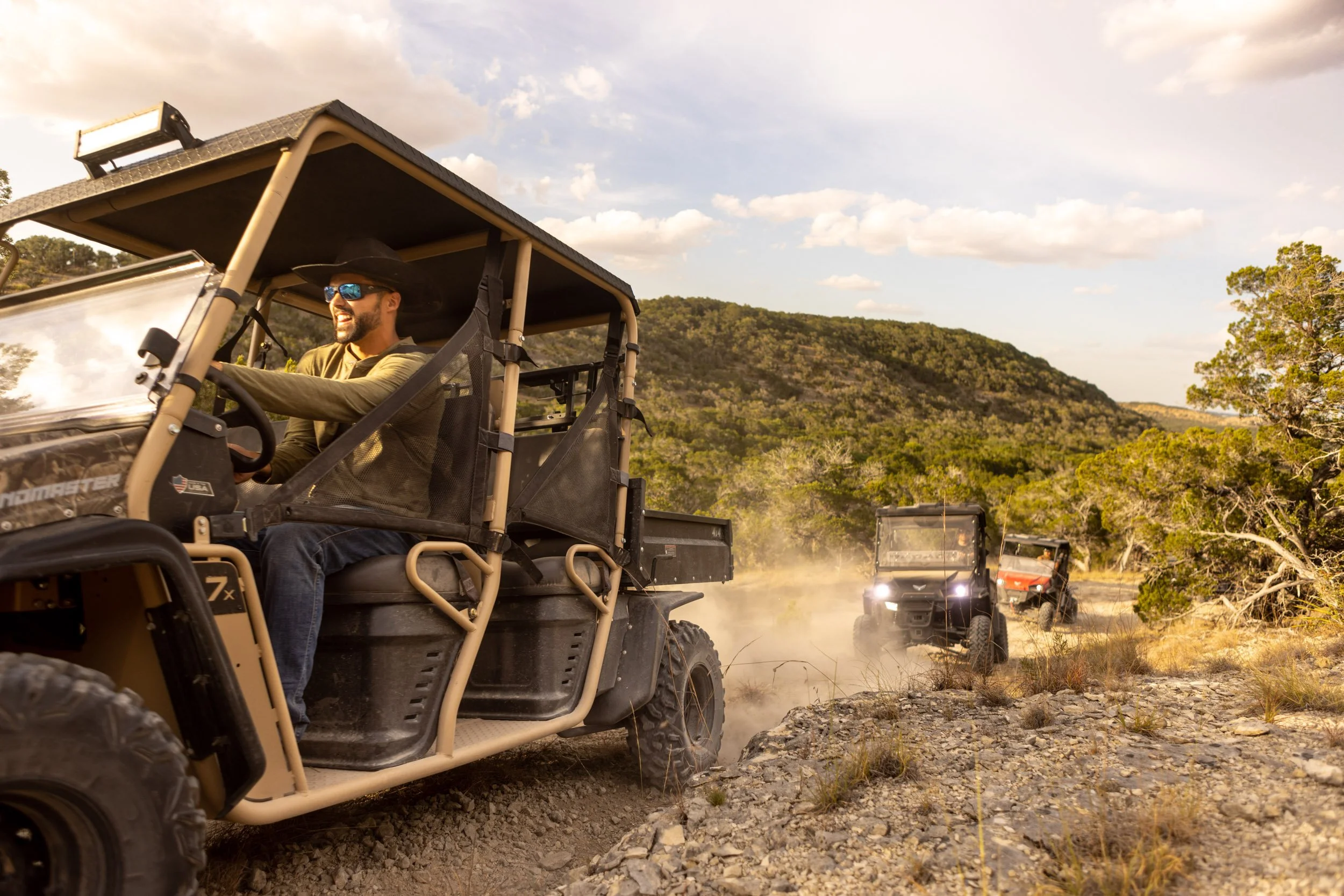 A man wearing sunglasses and a wide-brimmed hat is driving a beige utility vehicle through a dusty, rocky off-road landscape, with multiple similar vehicles following behind him. The background features forested hills, a partly cloudy sky, and a dry,