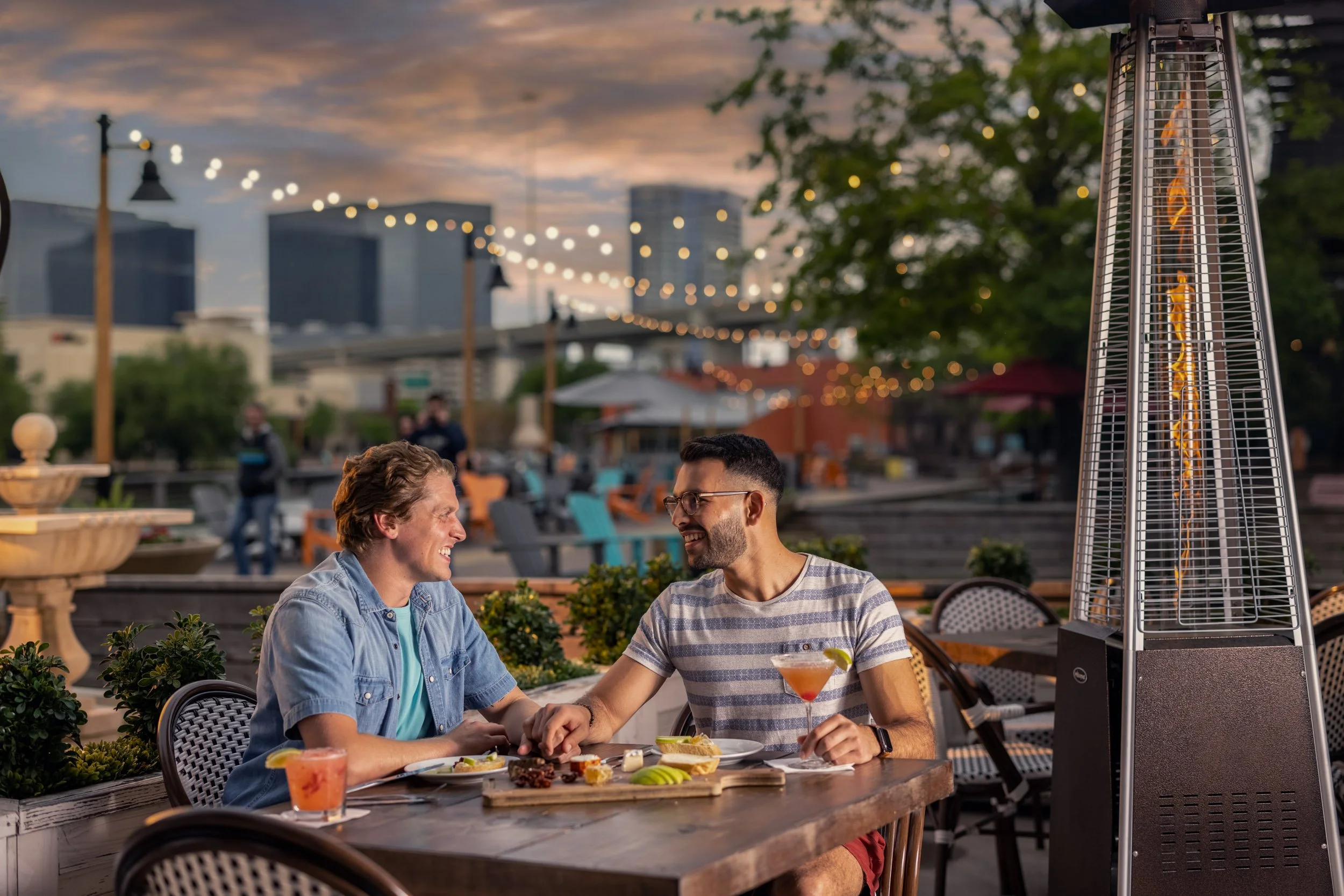 Two young men sitting at an outdoor restaurant table, smiling and holding hands, with a martini drink and food in front of them. Patio heater with glowing elements, string lights, and cityscape in the background at dusk.