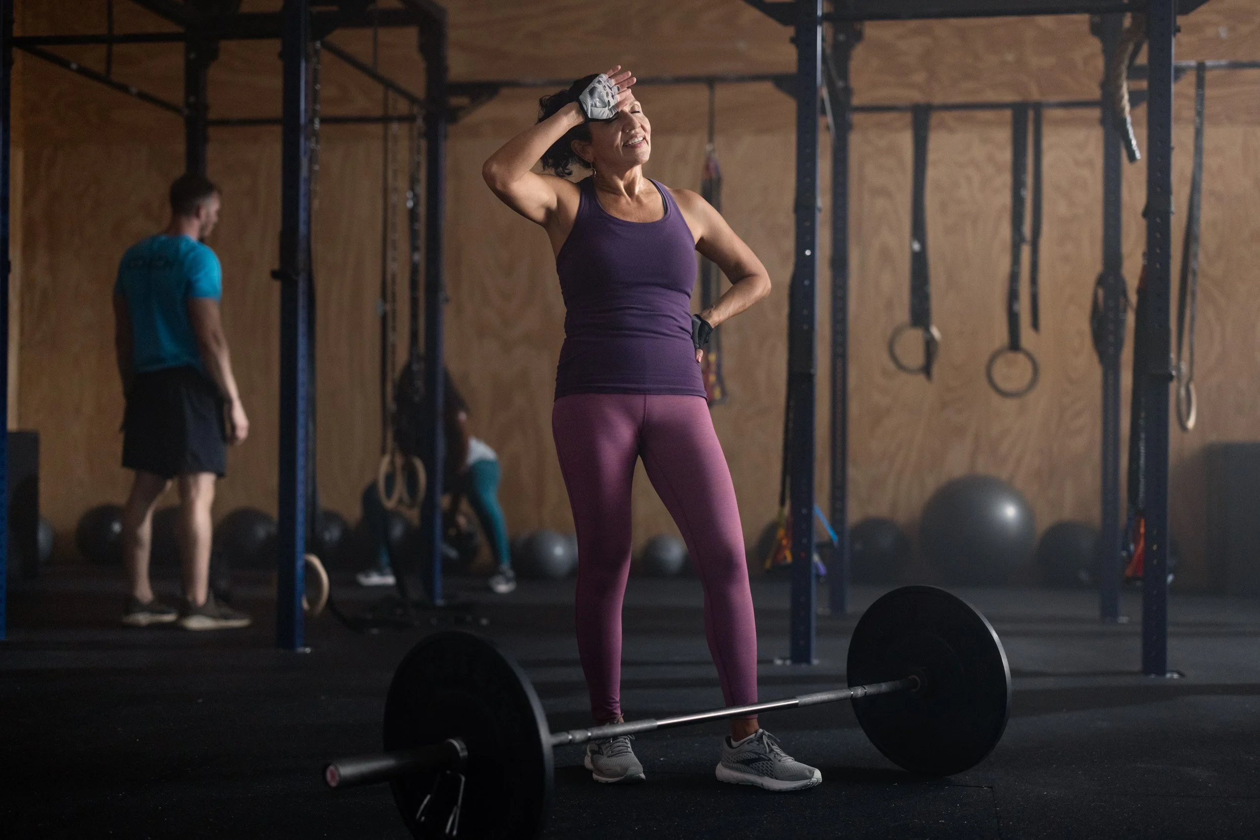A woman in workout clothing, smiling and wiping sweat from her forehead inside a gym, with gym equipment and other people exercising in the background.