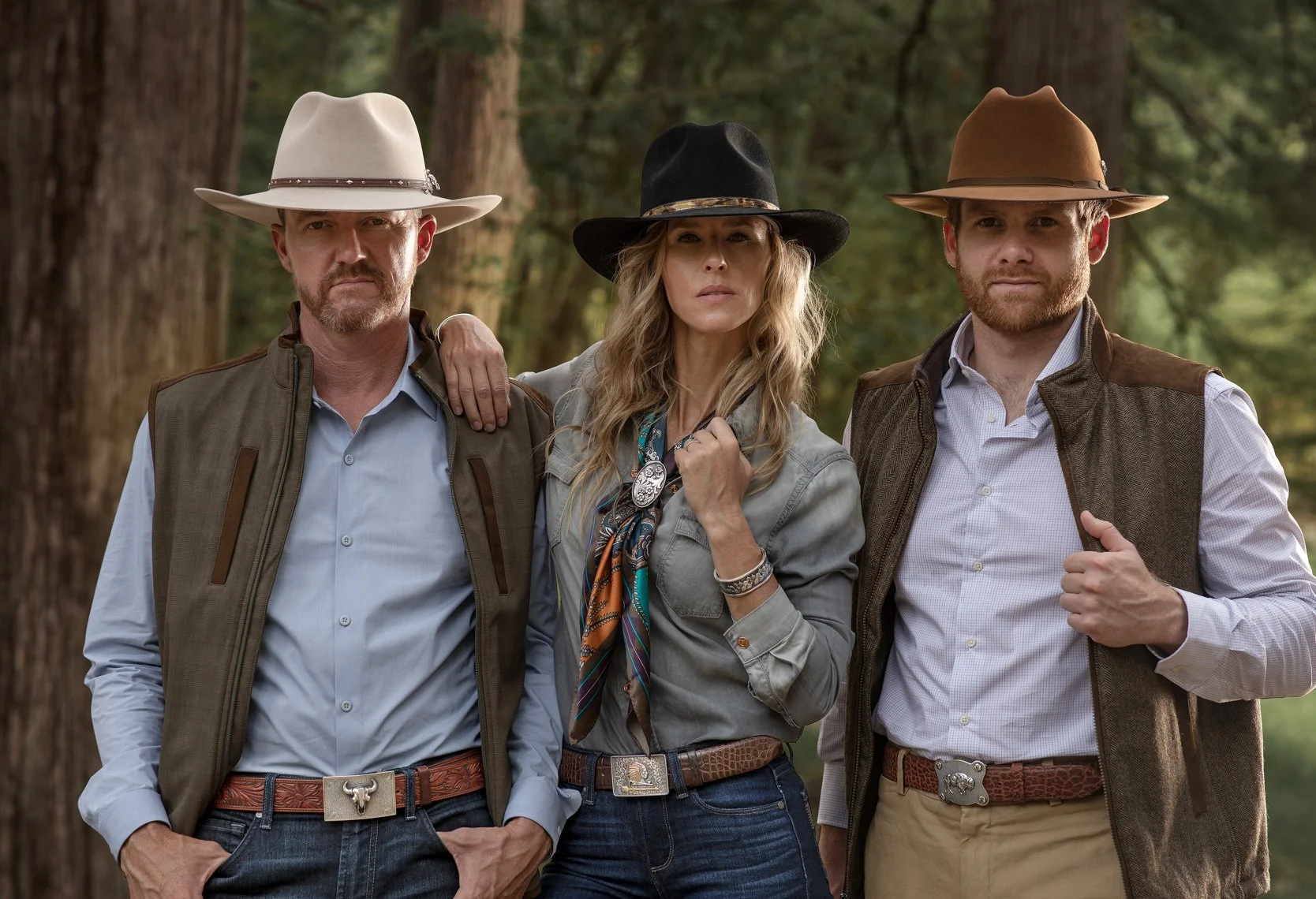 Three people in cowboy-style attire standing outdoors in a wooded area. The man on the left has a white cowboy hat, vest, and blue shirt. The woman in the middle has a black hat, long wavy hair, and a scarf around her neck. The man on the right has a