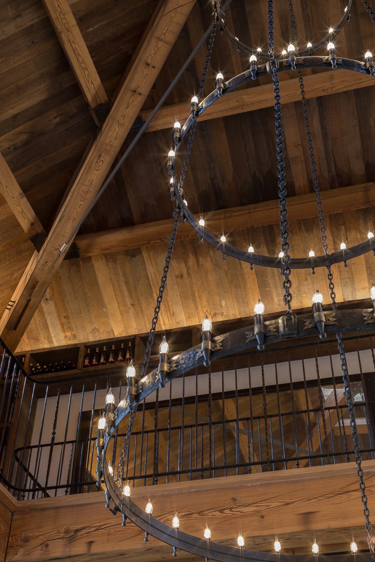 A rustic chandelier with two large circular metal rings and multiple small light bulbs, hanging from a wooden ceiling with exposed beams.
