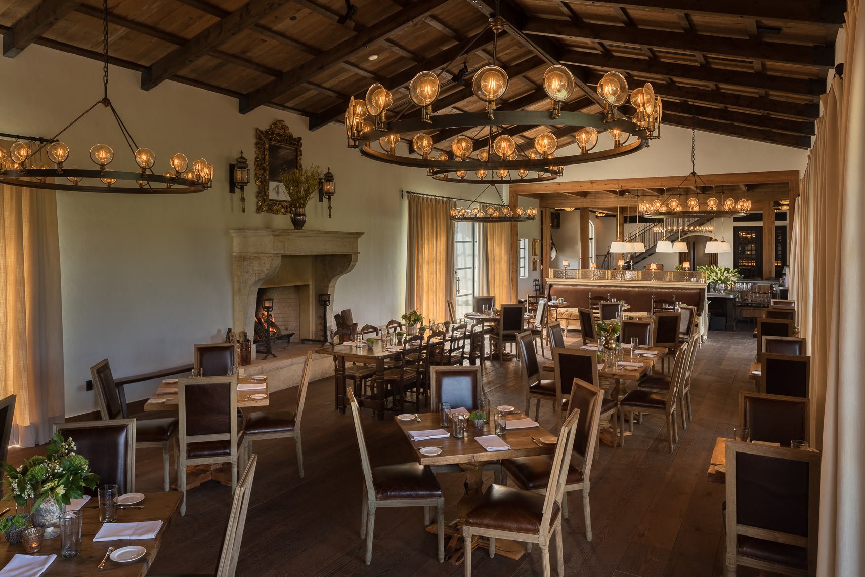 Interior of an elegant restaurant with wooden beams and chandeliers. Tables are set with plates, glasses, and napkins, surrounded by wooden chairs with leather cushions. A fireplace with a fire burning and decorative plants and lanterns are visible.