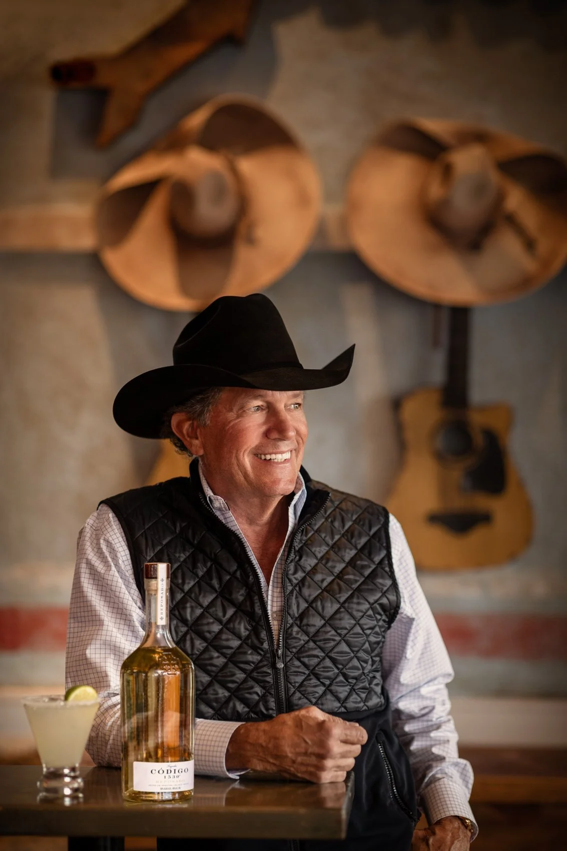 A smiling man in a black cowboy hat and a checkered shirt with a black quilted vest stands at a bar with a bottle of tequila and a lime wedge drink in front of him, against a background of mounted guitars and a wooden horse head.