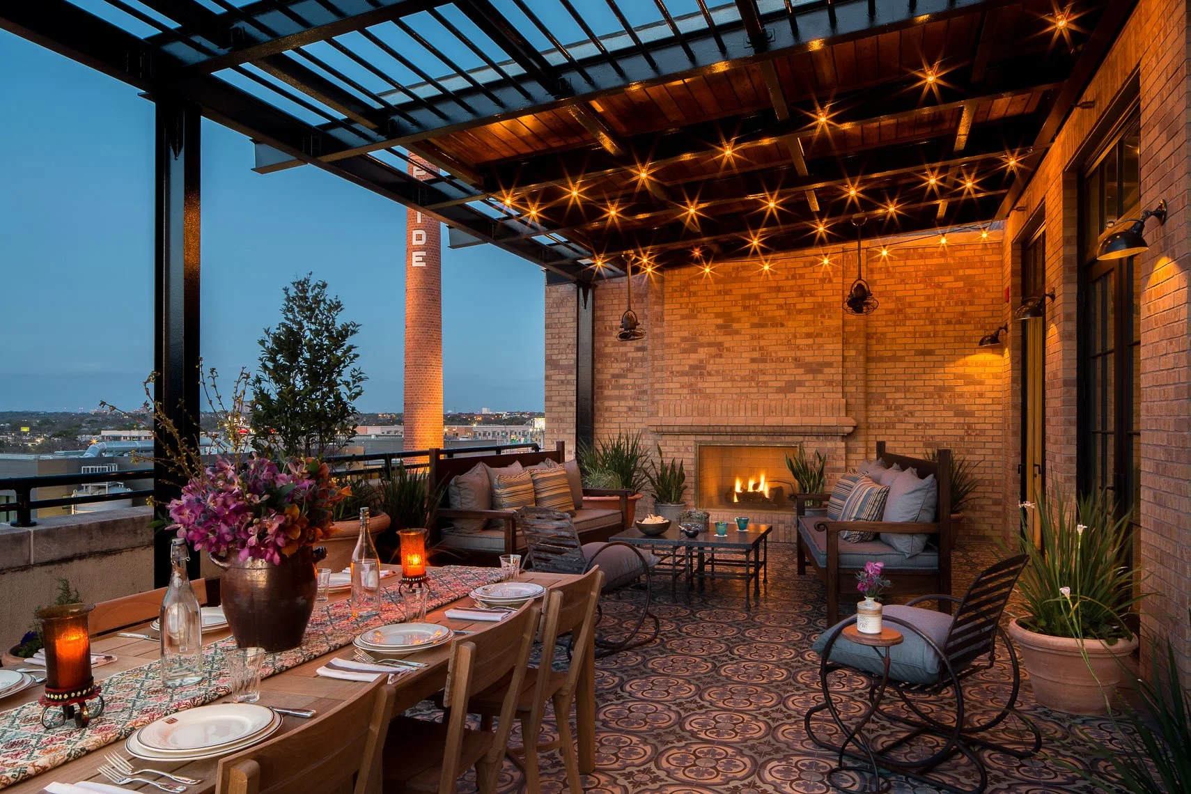 A cozy rooftop patio or lounge area during the evening with a fireplace, string lights, and outdoor furniture including a dining table with chairs and a seating area with sofas and armchairs. There are potted plants, flowers, and a cityscape view in 