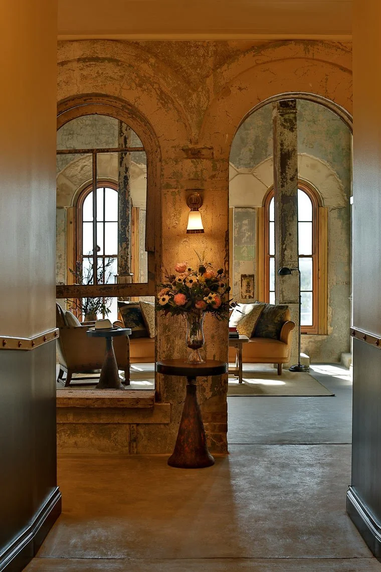 Interior of a cozy vintage living space with peeling paint walls, arched windows, and a vase of fresh flowers on a small table.