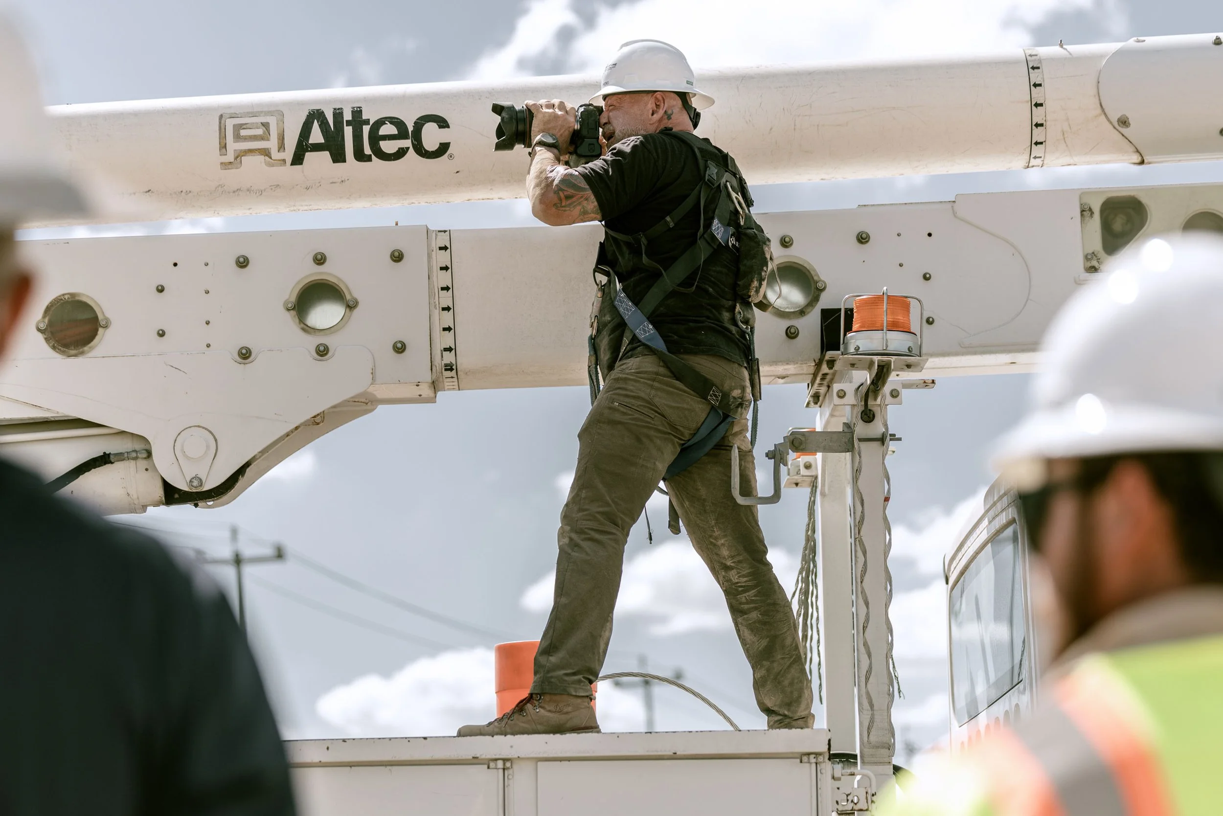 Worker wearing a helmet and harness taking a photo or looking through binoculars on a cherry picker lift, with a cloudy sky in the background.