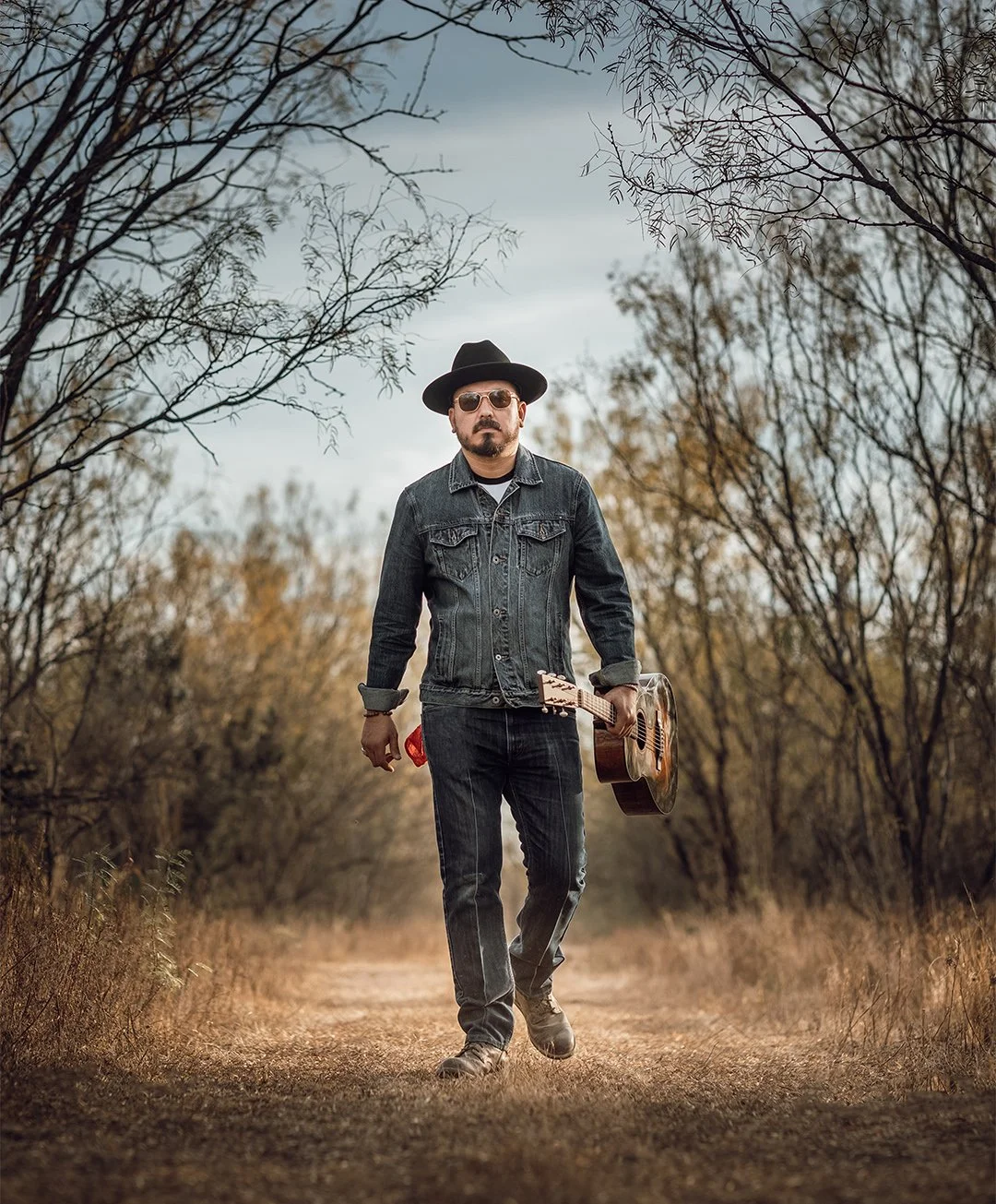 A man walking on a dirt path in a wooded area, holding an acoustic guitar in his right hand and wearing sunglasses, a denim jacket, a wide-brimmed hat, and jeans.