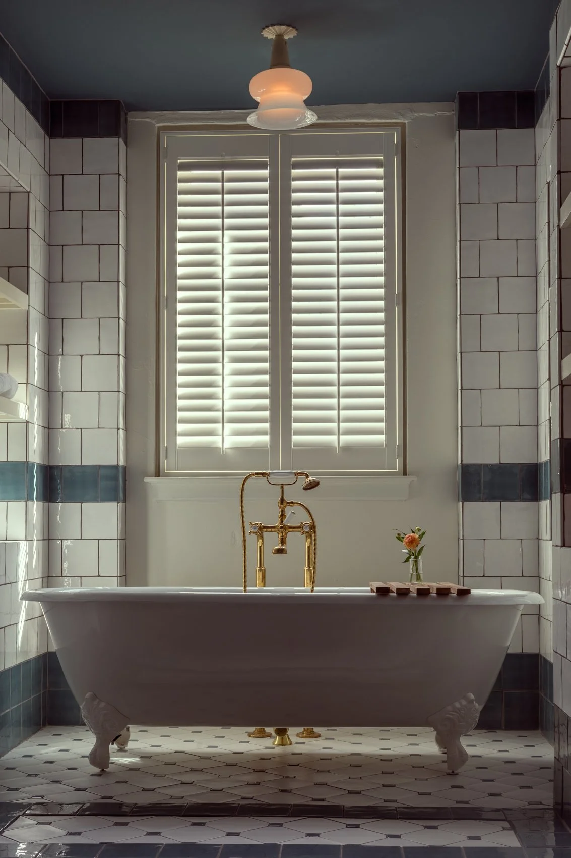 A classic white clawfoot bathtub with gold fixtures in a bathroom with white tiled walls and a window with closed shutter blinds.