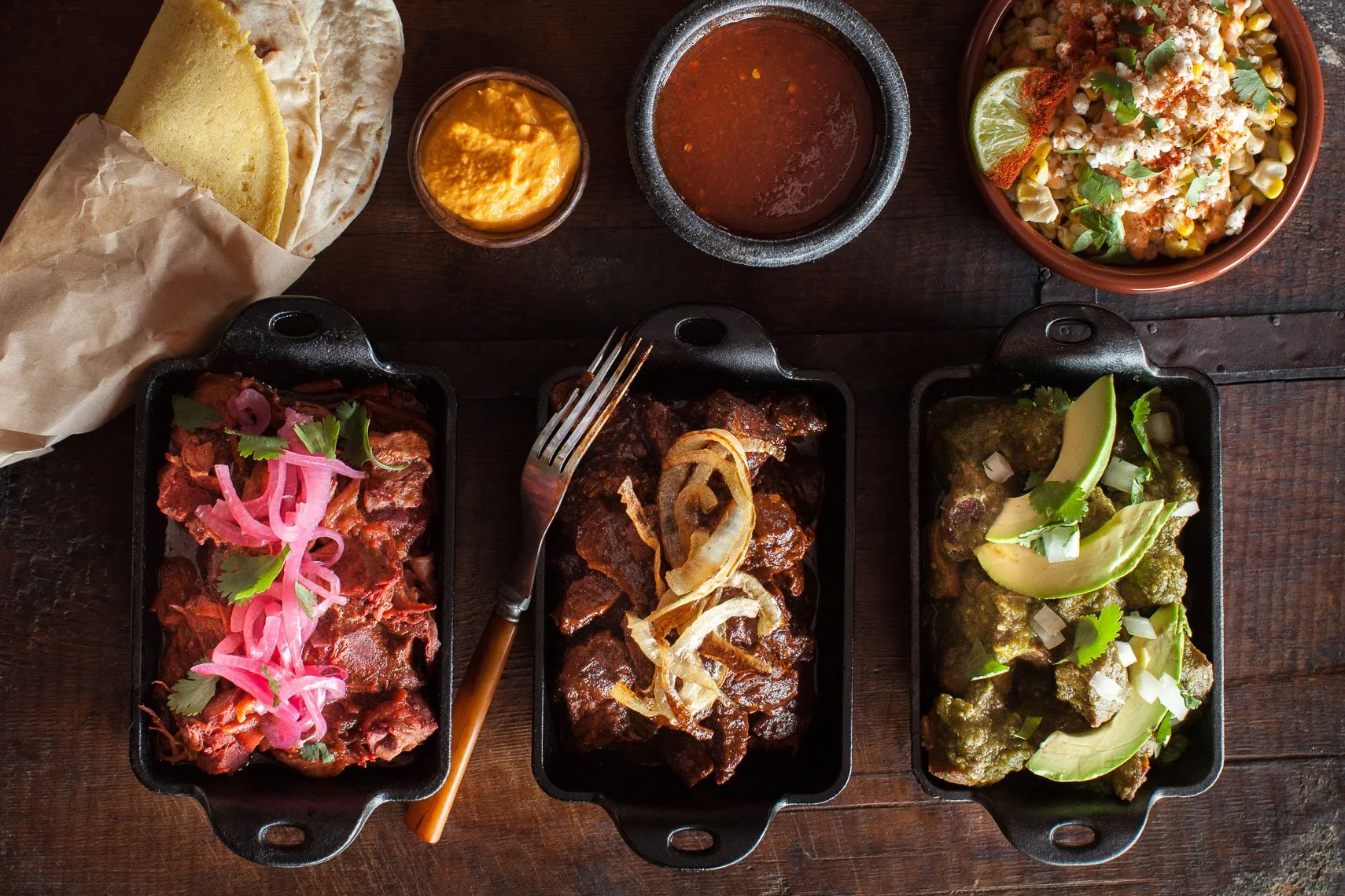 Three black rectangular dishes with Mexican meat dishes topped with fresh cilantro, onions, and pickled red onions, with cut lime slices and small bowls of sauces and drinks nearby on a wooden table.