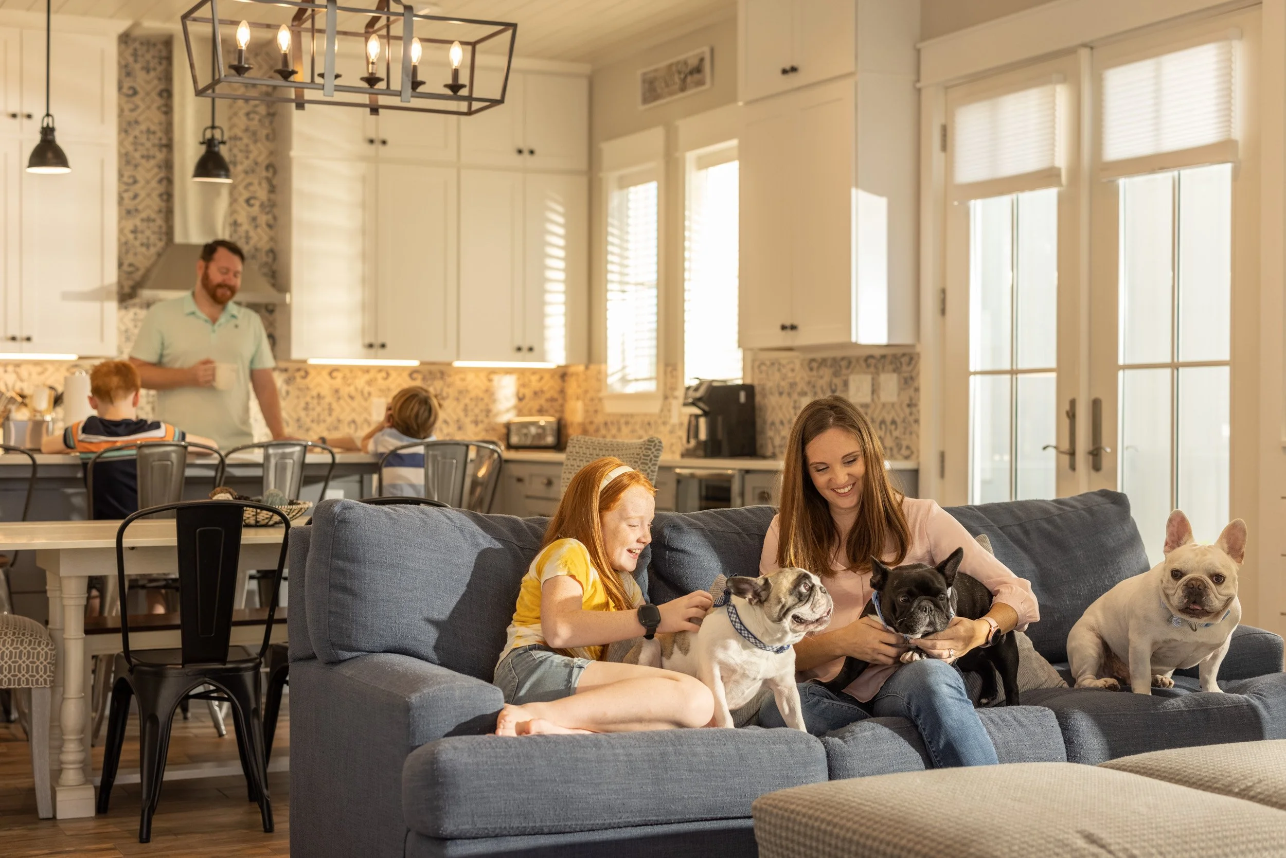 Family gathering in the living room with children, woman, and dogs, while man prepares coffee in the kitchen.