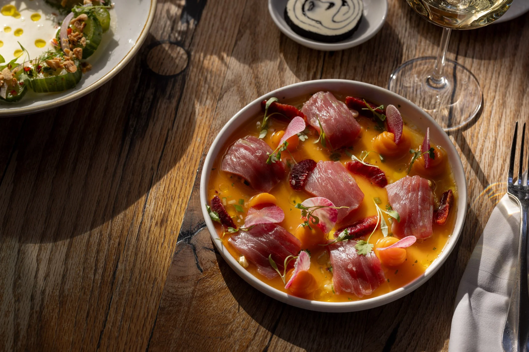 A bowl of scrambled eggs garnished with slices of prosciutto and edible flowers. In the background, there is a plate of sliced cucumbers topped with herbs and nuts, and a small bowl of black and white swirl dip. A glass of white wine is on the wooden