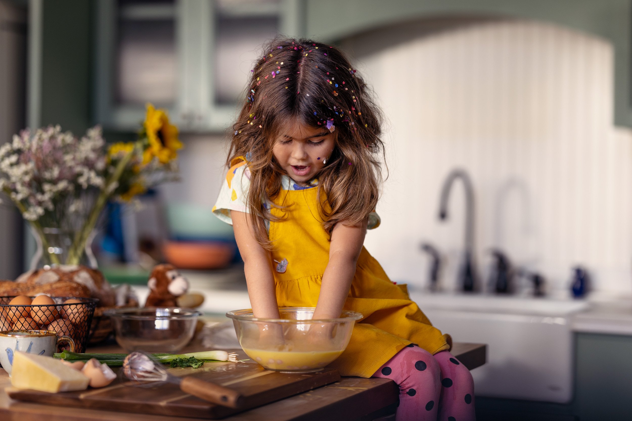 A young girl with long brown hair covered in colorful confetti, wearing a yellow dress and pink polka dot tights, is mixing ingredients in a glass bowl in a kitchen.