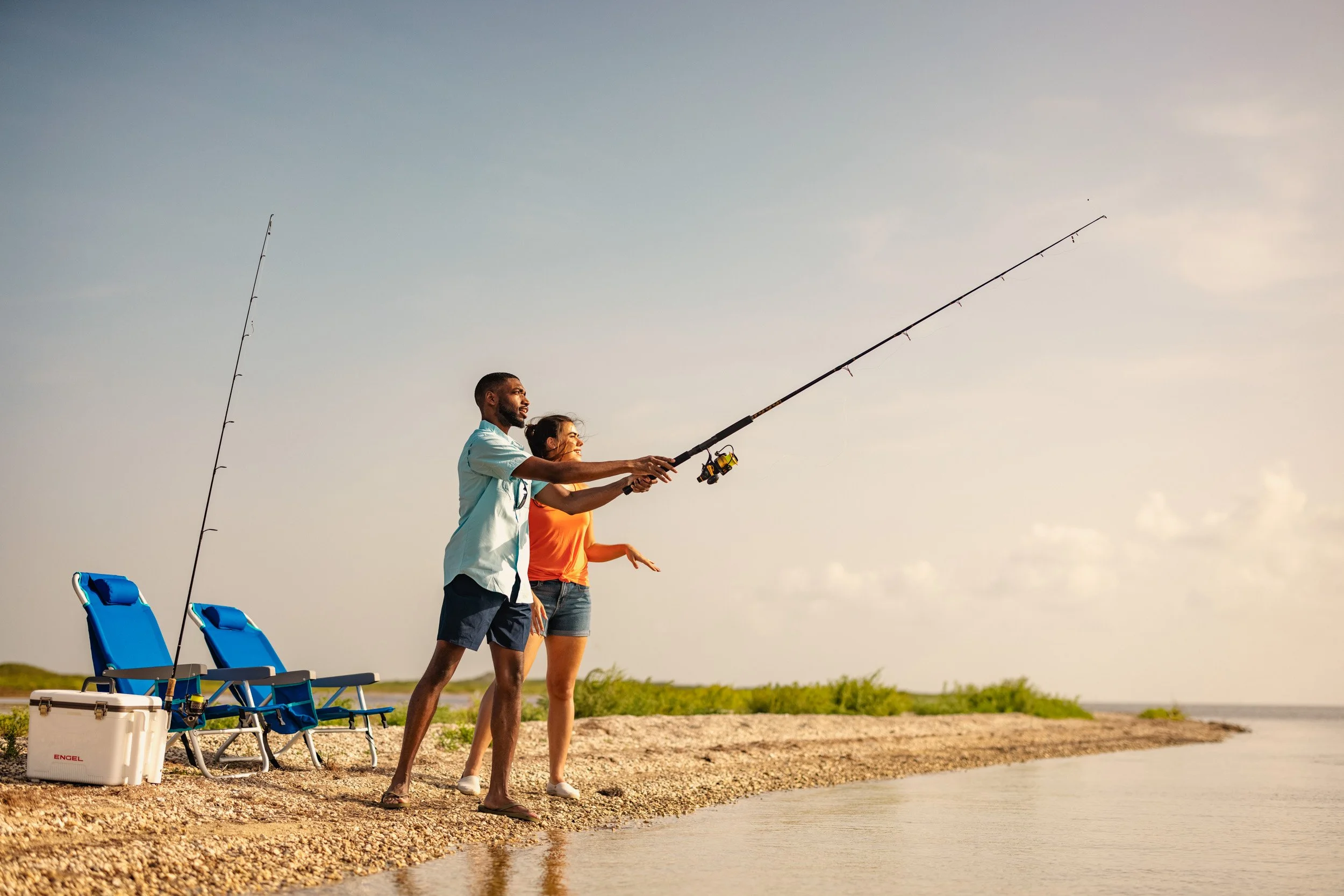 A man and woman fishing by the water on a beach at sunset, with fishing gear and chairs nearby.