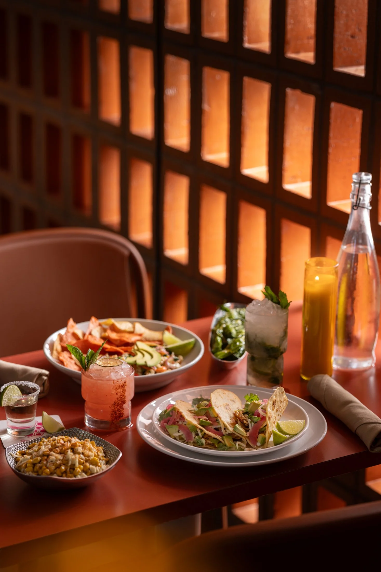 A dining table with various Mexican dishes including tacos, a bowl of corn salad, and drinks such as margaritas and bottled beverages. The background features a decorative brick wall with square compartments.