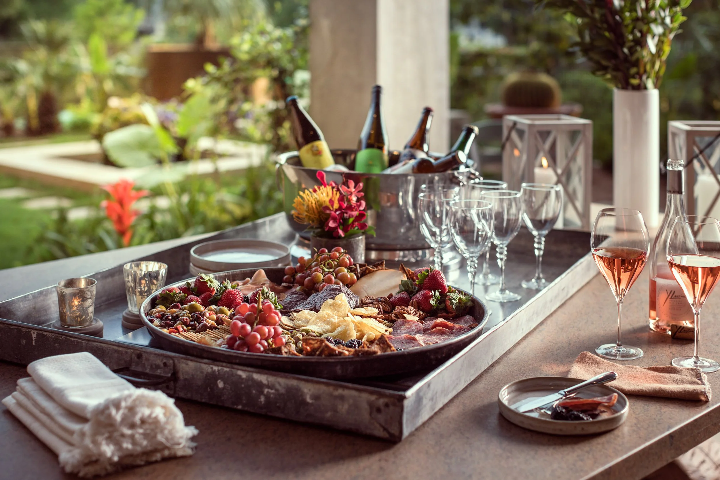A table set for a meal with a tray of assorted fruits, cheese, and cured meats, surrounded by wine glasses and candles, with a vase of purple flowers and a garden in the background.