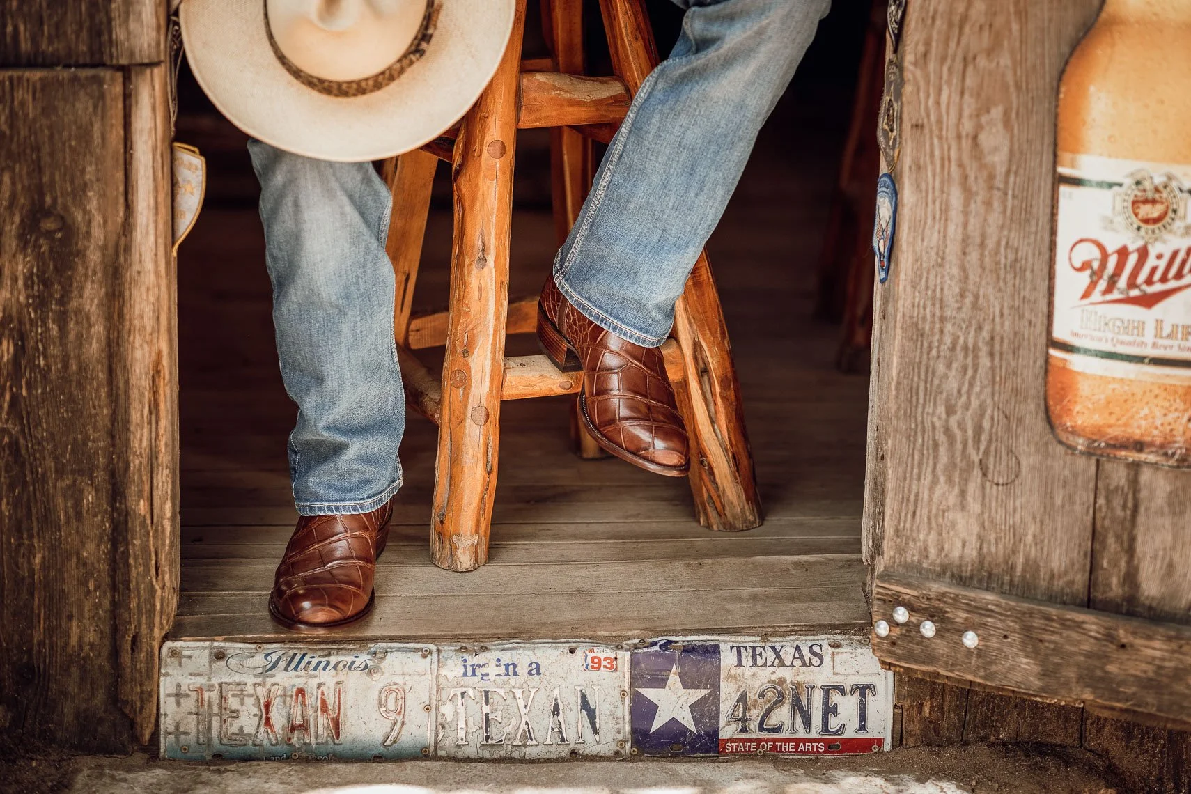 A person sitting on a rustic wooden bar stool wearing jeans and brown cowboy boots, holding a cowboy hat, with a wooden bar area and Texas and Illinois license plates visible underneath.