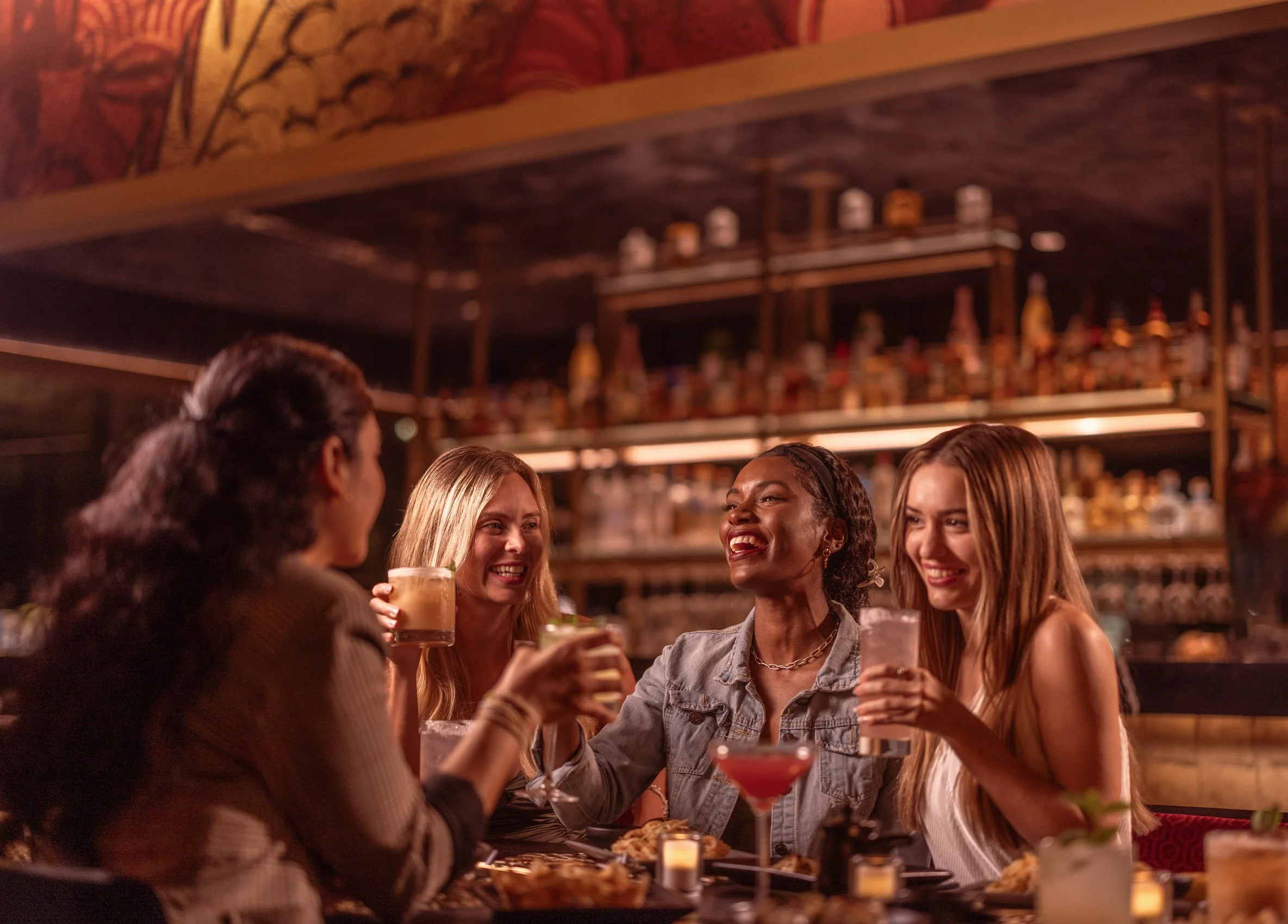 Group of six women enjoying drinks and conversation in a dimly lit, cozy bar or restaurant.