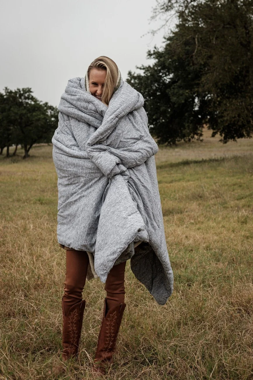 Woman standing outdoors in a field, wrapped in a large gray blanket, wearing brown boots, and smiling.