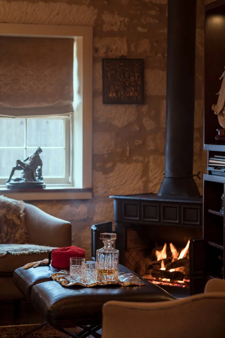 A cozy living room with a lit fireplace, a window with blinds, a statue on the window sill, a sofa with a fur throw, a tray with glassware and a decanter, and a bookshelf.