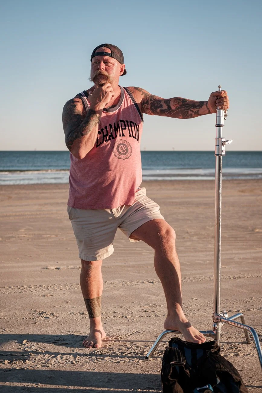 A man with tattoos, a beard, and a backwards cap standing on a sandy beach at sunset, holding a lighting stand and looking thoughtful.