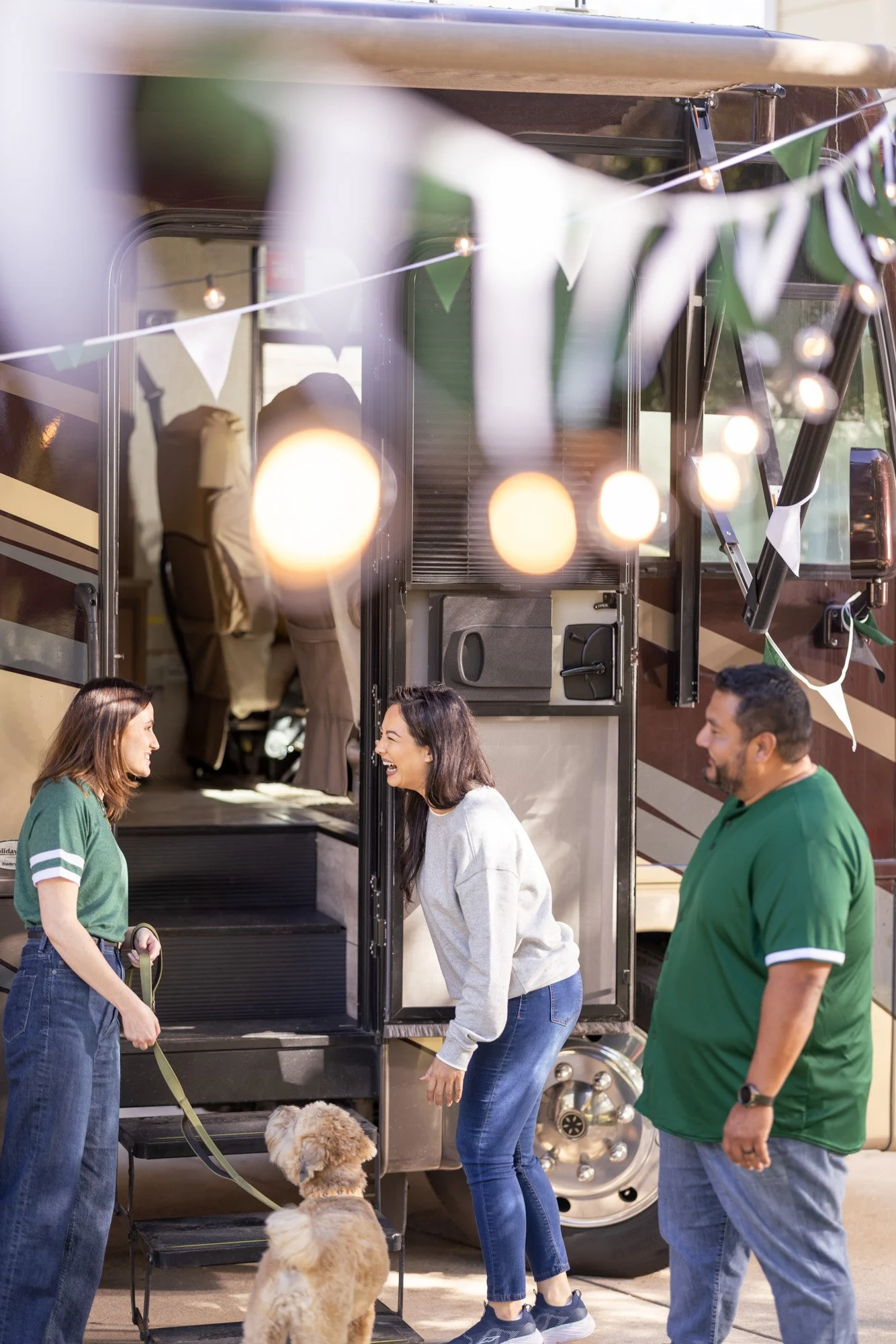 People interacting outside a food truck, with two women shaking hands, a woman in a white sweater, a man in a green shirt, and dogs on leashes nearby, with festive bunting and string lights overhead.
