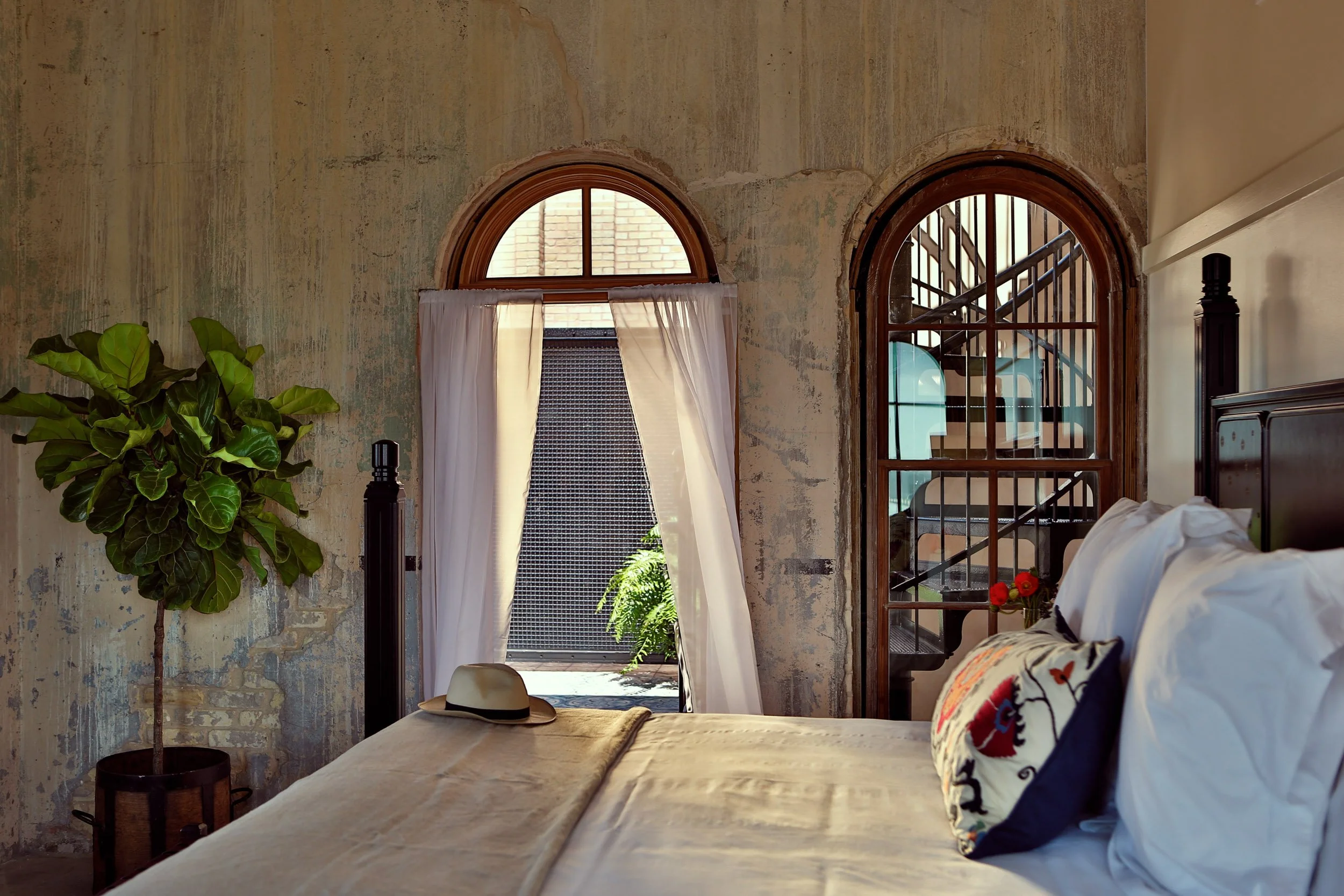 A bedroom with rustic textured walls, two arched windows with sheer white curtains, a large green plant, a hat on the bed, and a black bed frame with a white comforter and colorful pillows.