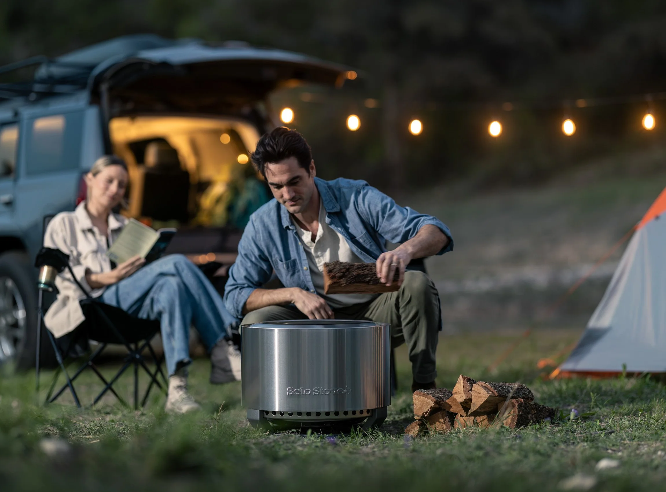 Two people camping outdoors during the evening, sitting by a fire pit with firewood nearby, with a vehicle and string lights in the background.