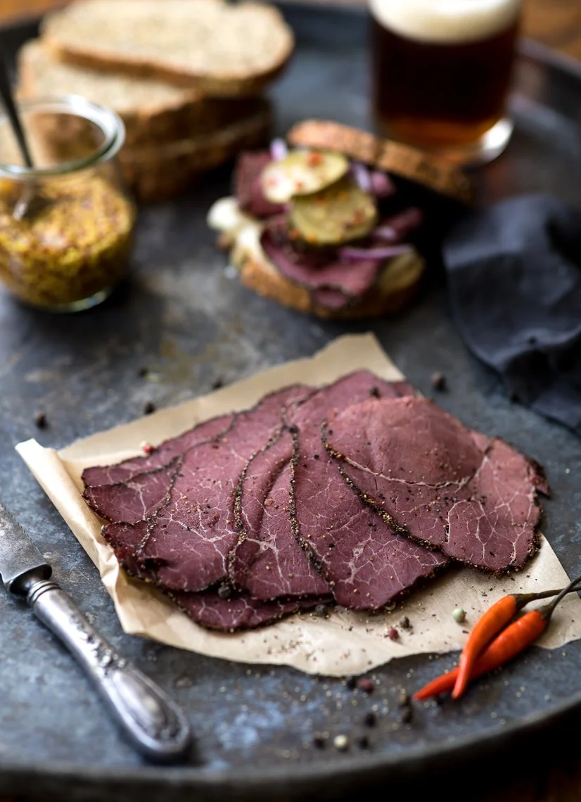 Sliced roast beef seasoned with cracked black pepper, served on parchment paper with fresh carrots nearby, and slices of bread and a jar of mustard in the background.