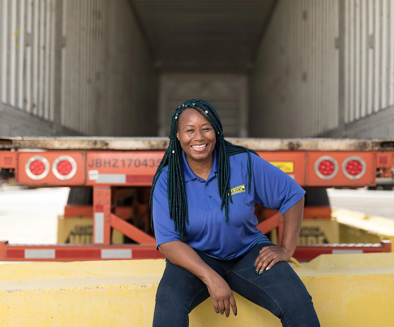 A woman with braided hair, smiling, wearing a blue uniform shirt and sitting on a yellow curb in front of a semi-truck trailer.