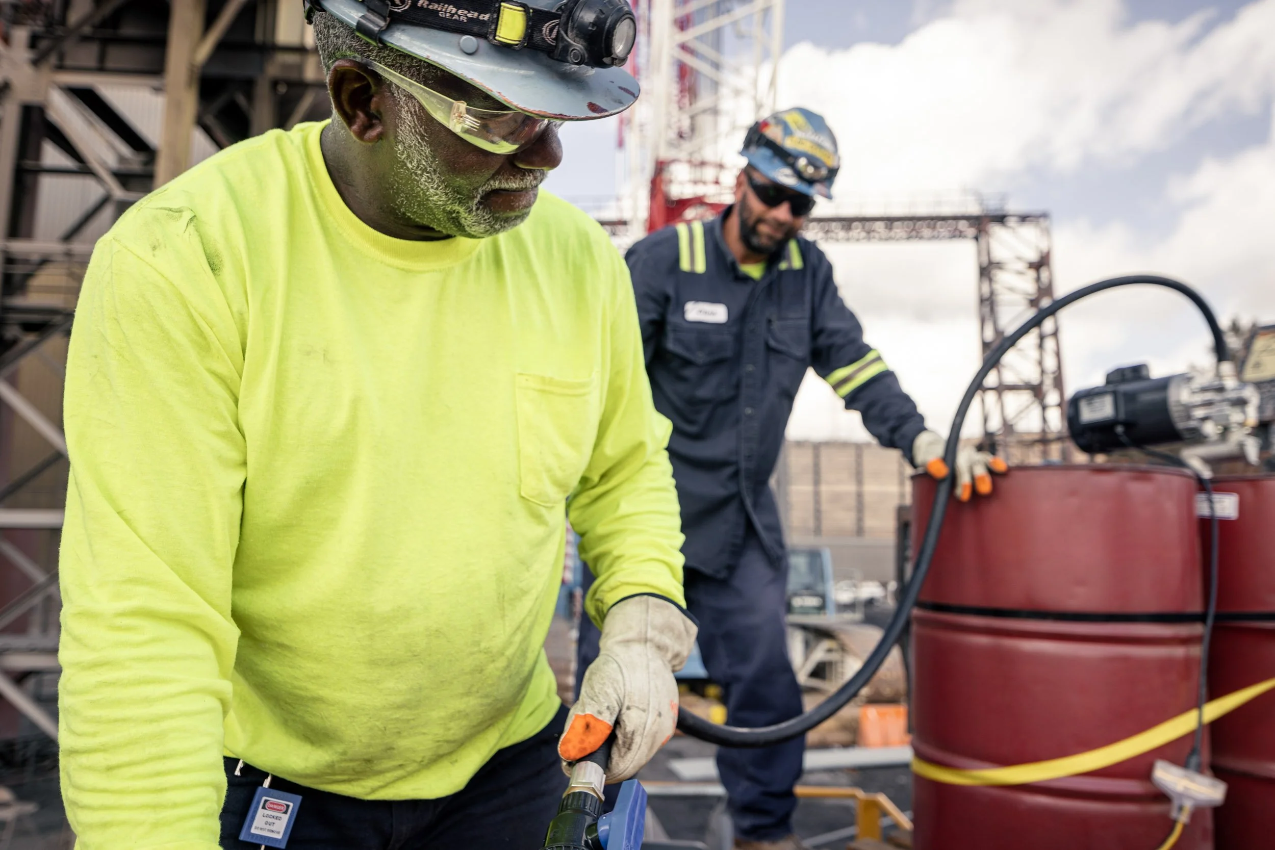 Two construction workers at a construction site, one wearing a bright yellow shirt, safety helmet, and goggles, using a power drill, with scaffolding and metal structures in the background.