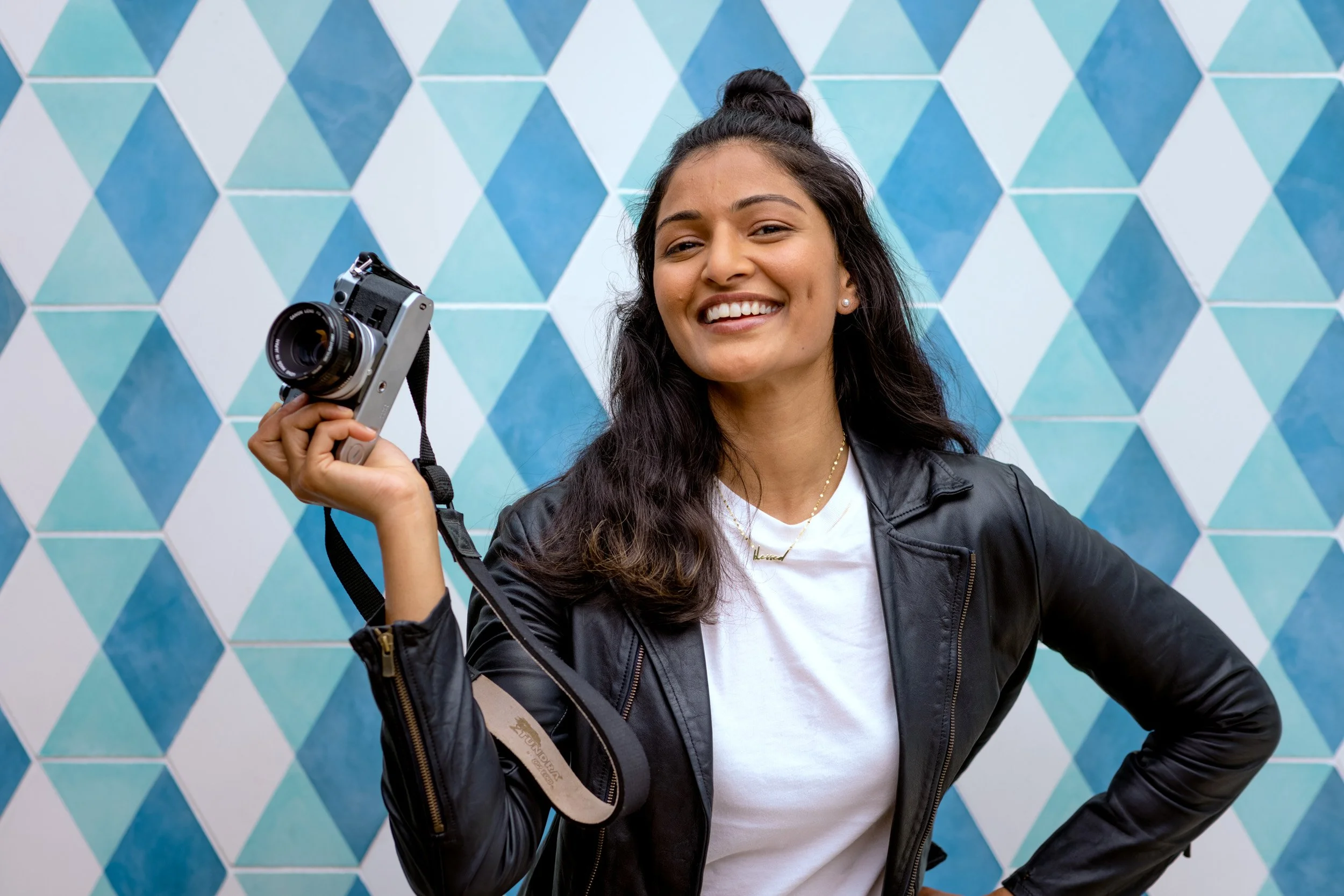 A young woman with long dark hair in a half-up hairstyle, wearing a black leather jacket, white t-shirt, and pearl earrings, smiling while holding a camera in front of a geometric blue and teal patterned wall.